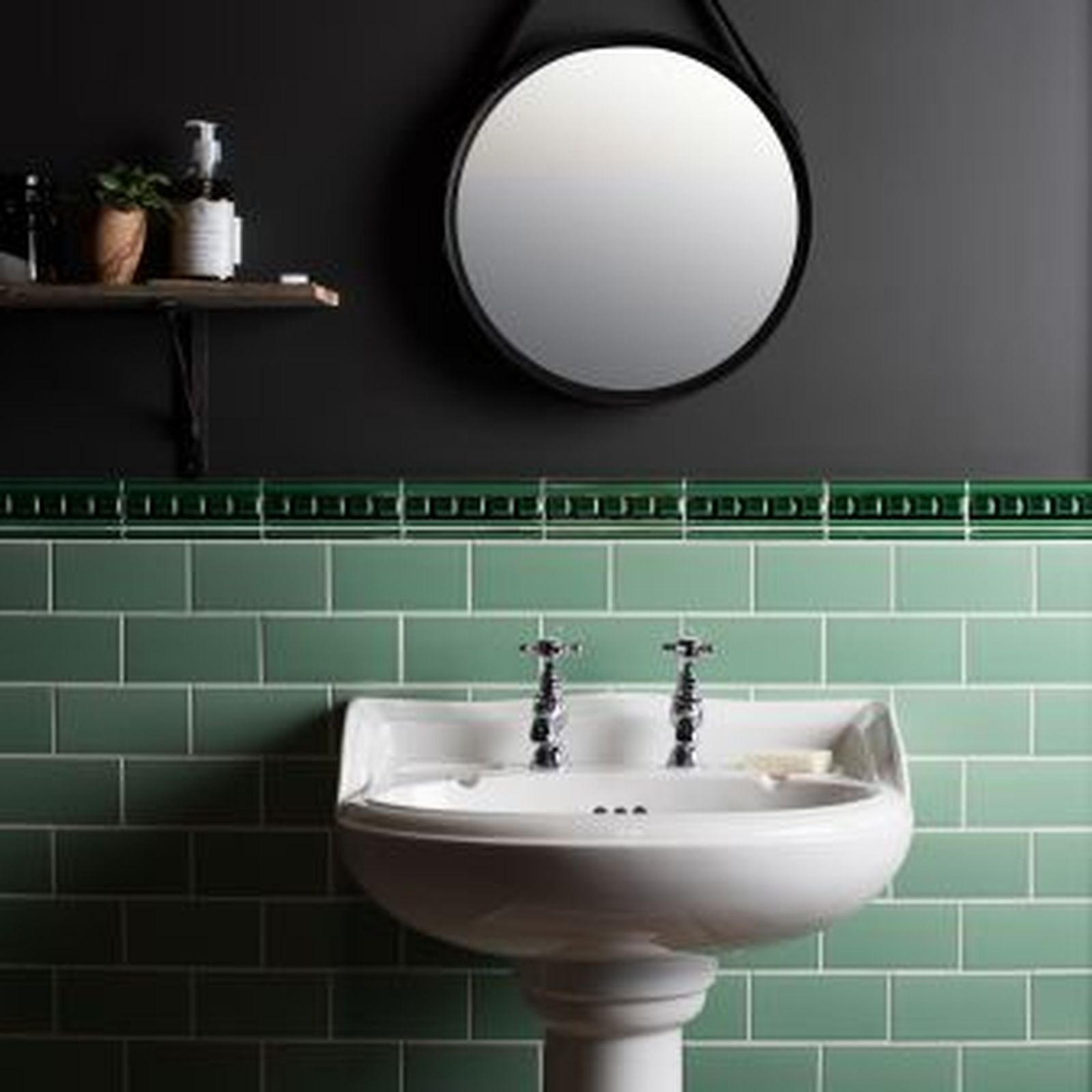 Bathroom with green tiled walls, white pedestal sink, round mirror, and shelf with plants and toiletries.