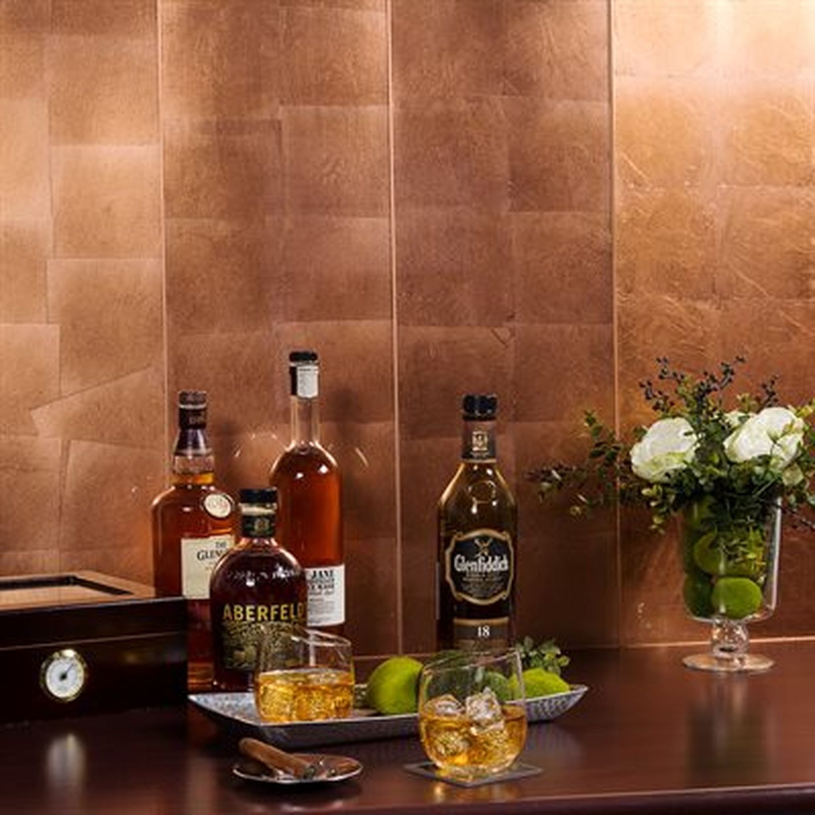 Bar setup with bottles, glasses of whiskey, limes, flowers, and copper-toned wall.