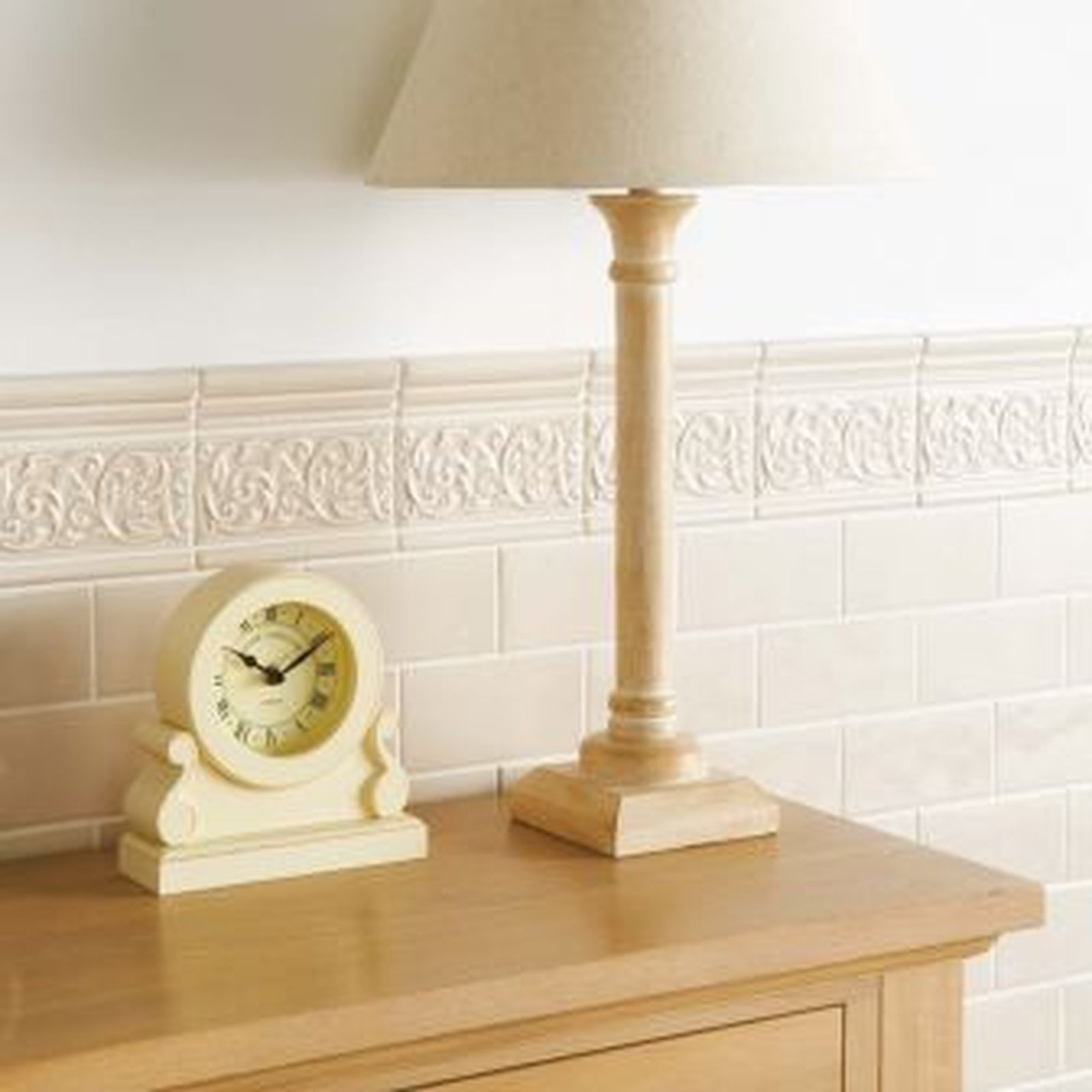 A cream-colored desk lamp and clock sit on a wooden table against a tiled wall.