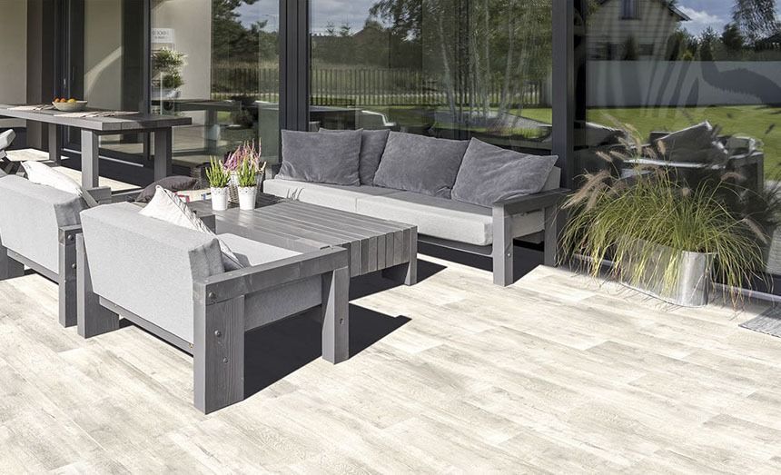 Patio furniture on light wood deck: gray sofa, chairs, table, plants, and large glass windows.