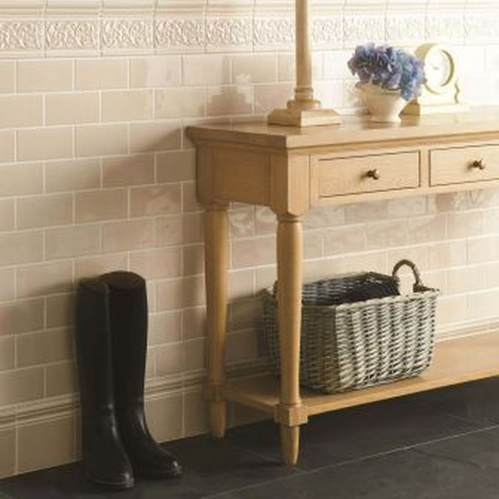Tan tiled wall with wood console table, basket, boots, lamp, and clock.