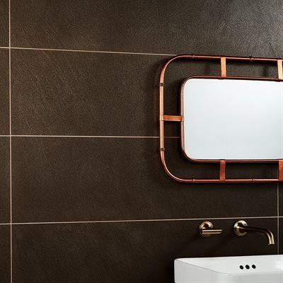 Dark brown tiled bathroom wall with copper-framed mirror and faucet over a white sink.