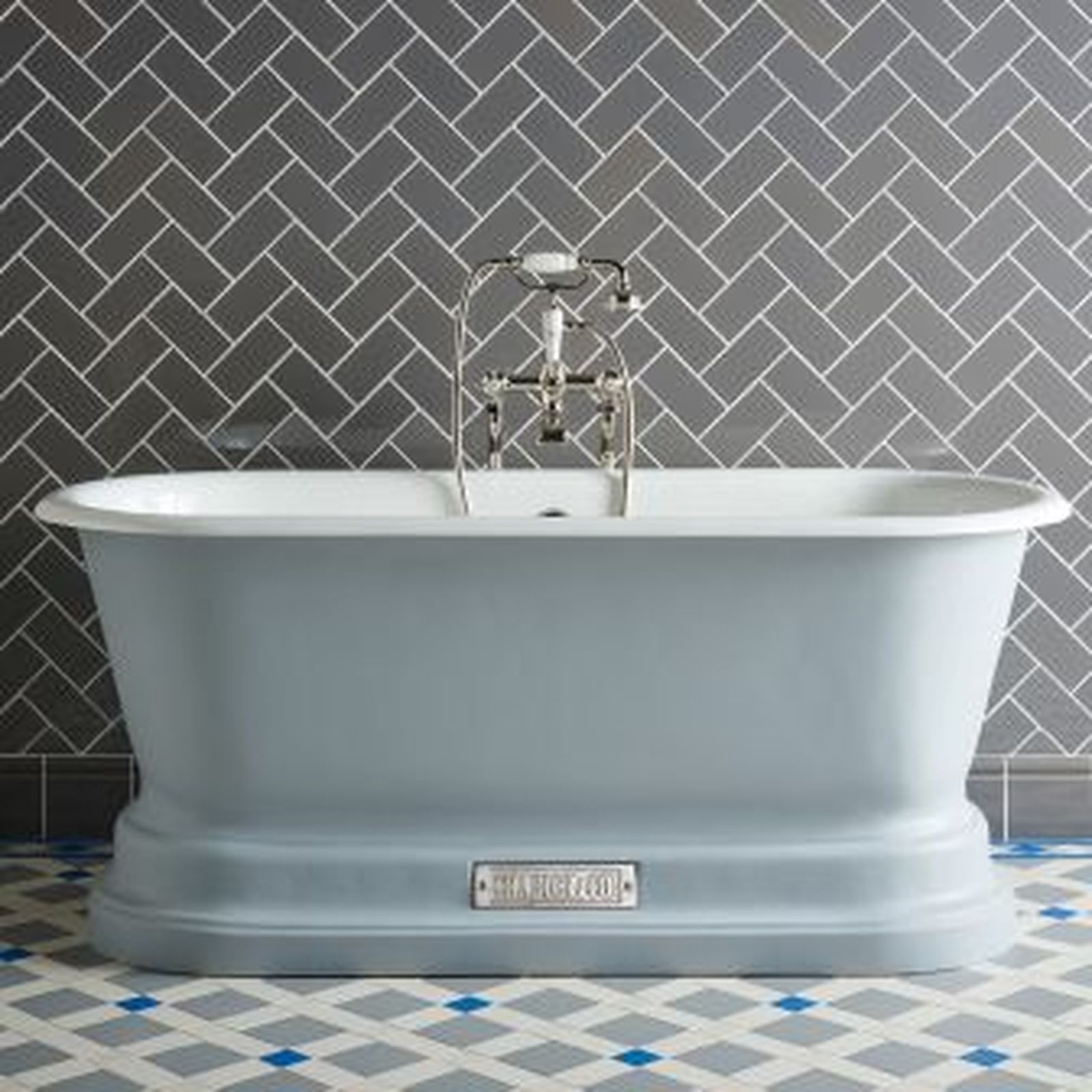 A light blue, freestanding bathtub with silver fixtures in front of a gray herringbone tile wall.