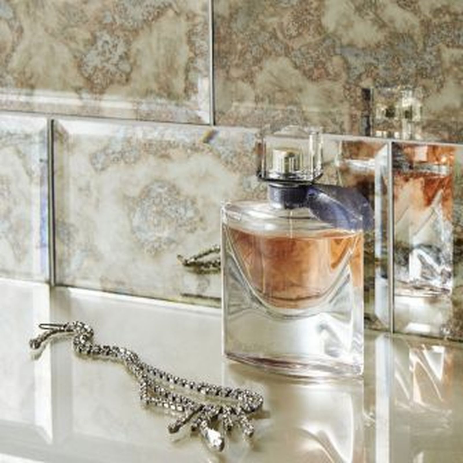 Perfume bottles and jewelry on a mirrored surface, against a textured wall.