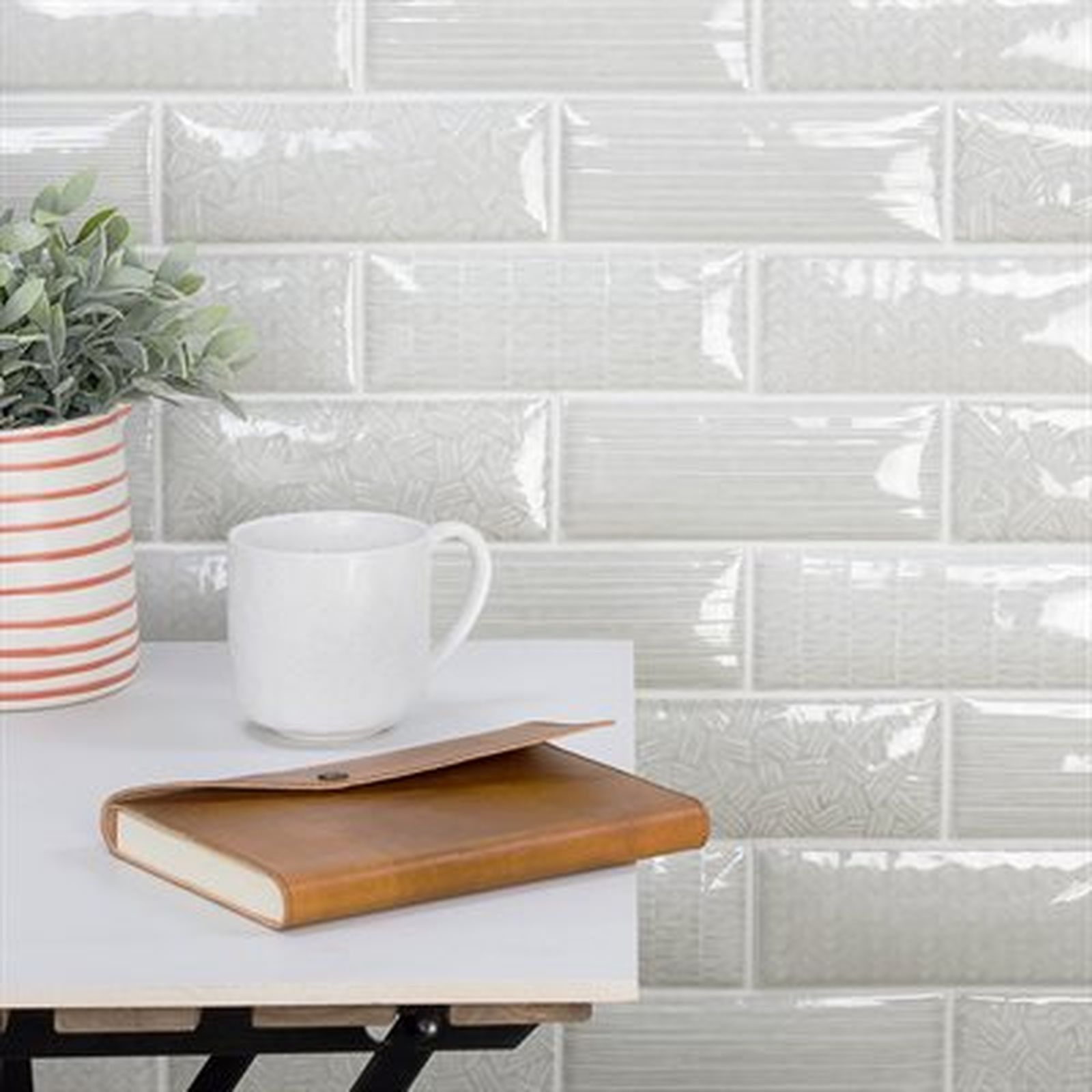 A white mug, brown notebook, and potted plant on a white table against a tiled wall.