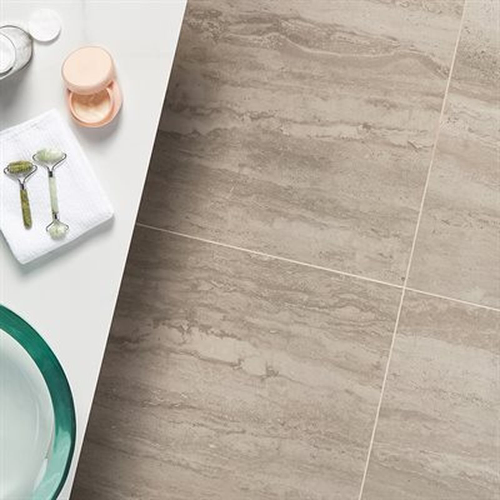 Bathroom tile floor with spa items on countertop: face roller, powder compact, skincare products, and a towel.
