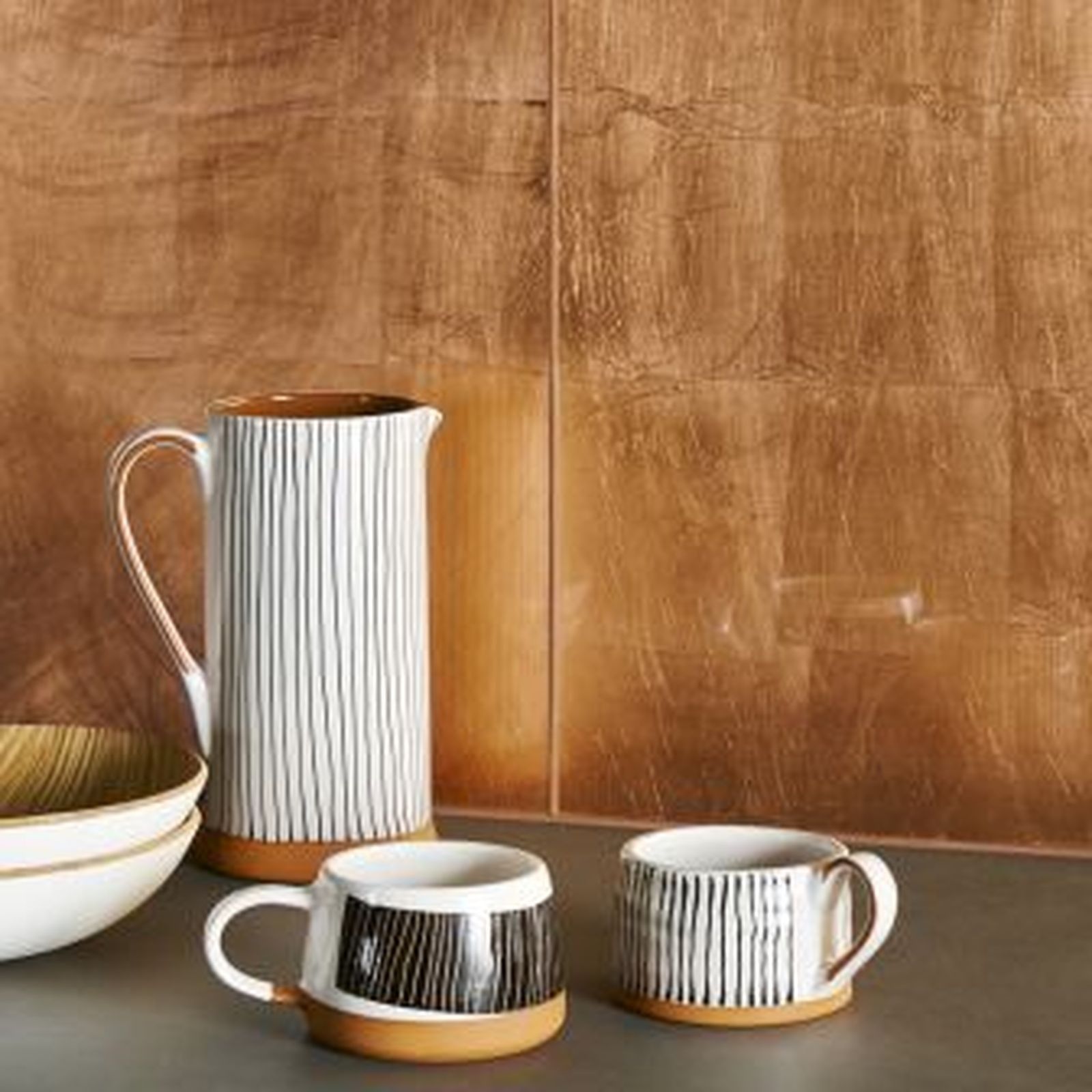 Ceramic pitcher, mugs, and bowl on a gray surface against a copper-colored wall.
