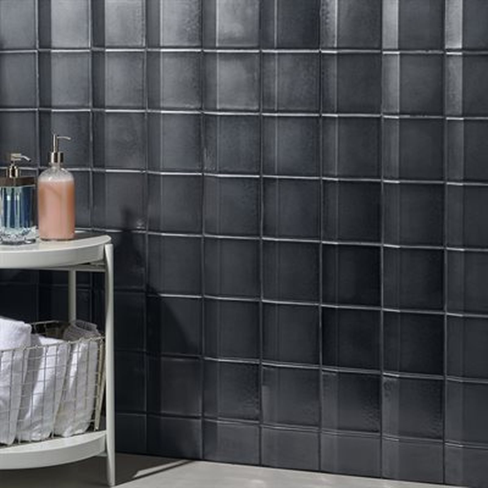 Black square tile wall with toiletries on a side table and a basket of towels.
