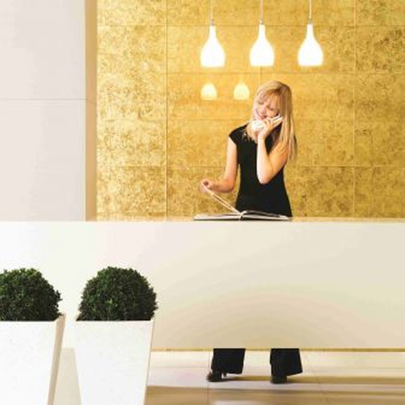 Woman at a cream-colored reception desk, talking on the phone. Golden wall behind. Two potted plants in white planters.