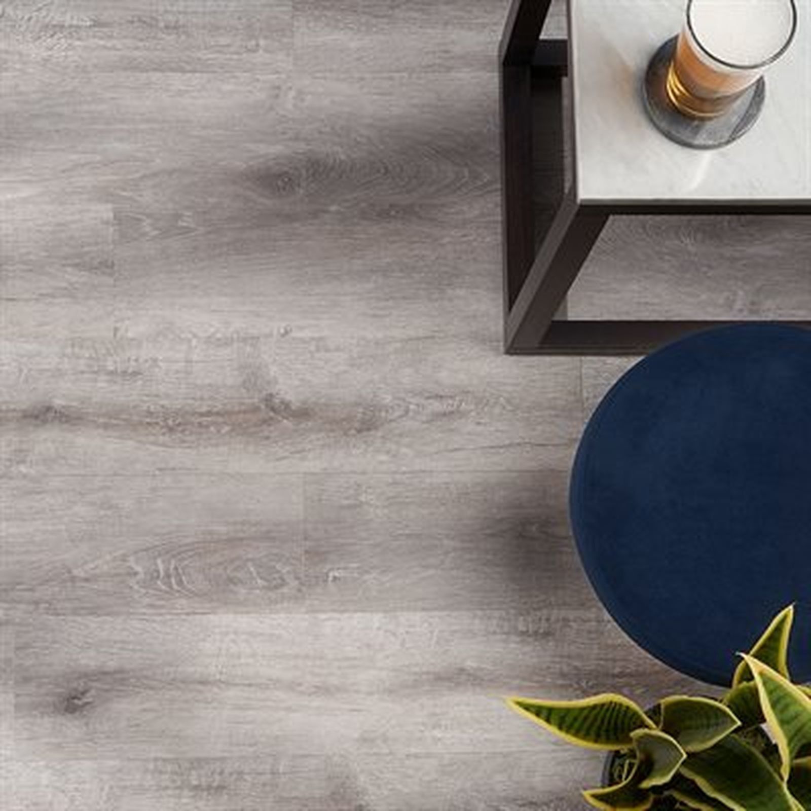 Gray wood-look flooring with a modern table holding a drink, a blue ottoman, and a plant.
