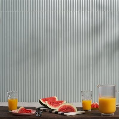 Watermelon slices, orange juice, and glasses on a wooden table in front of a ribbed, pale green wall.