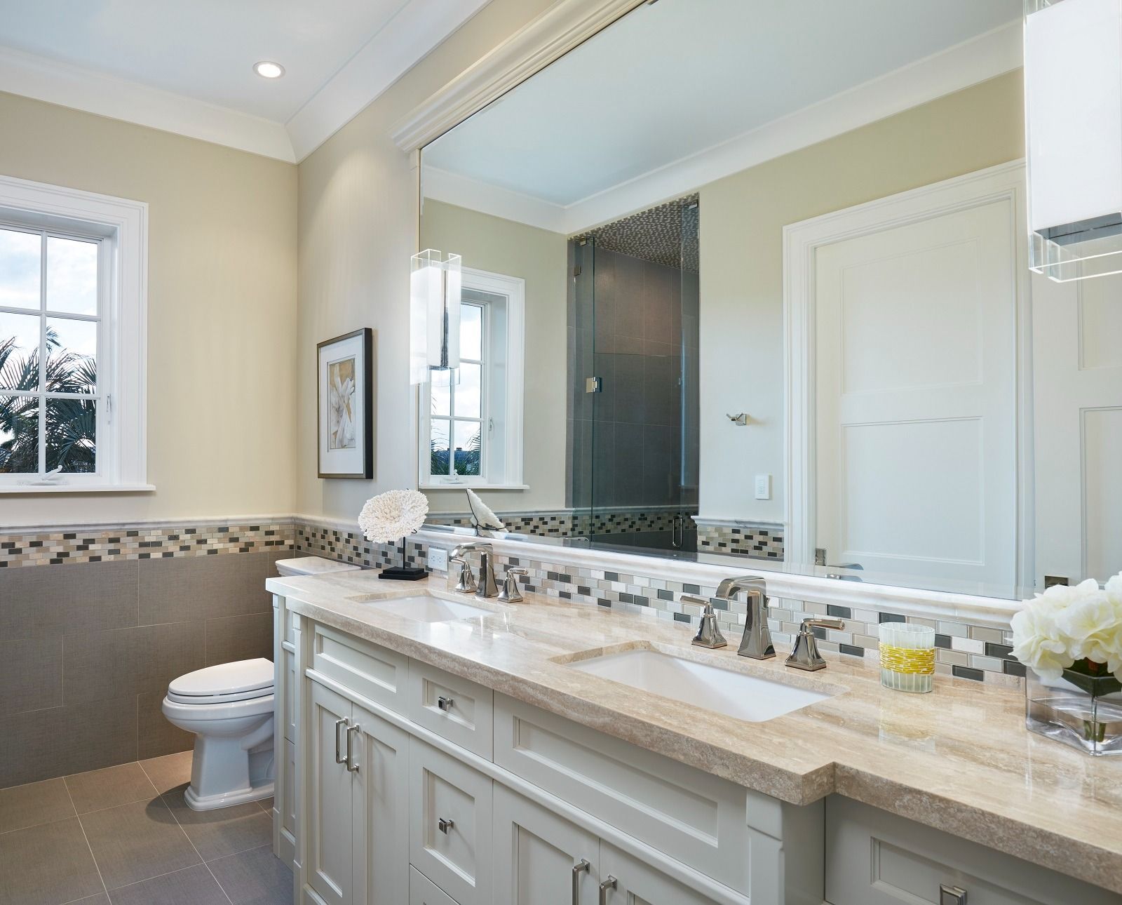 Bathroom with a large mirror, white cabinets, beige countertops, and a window with a view of trees.