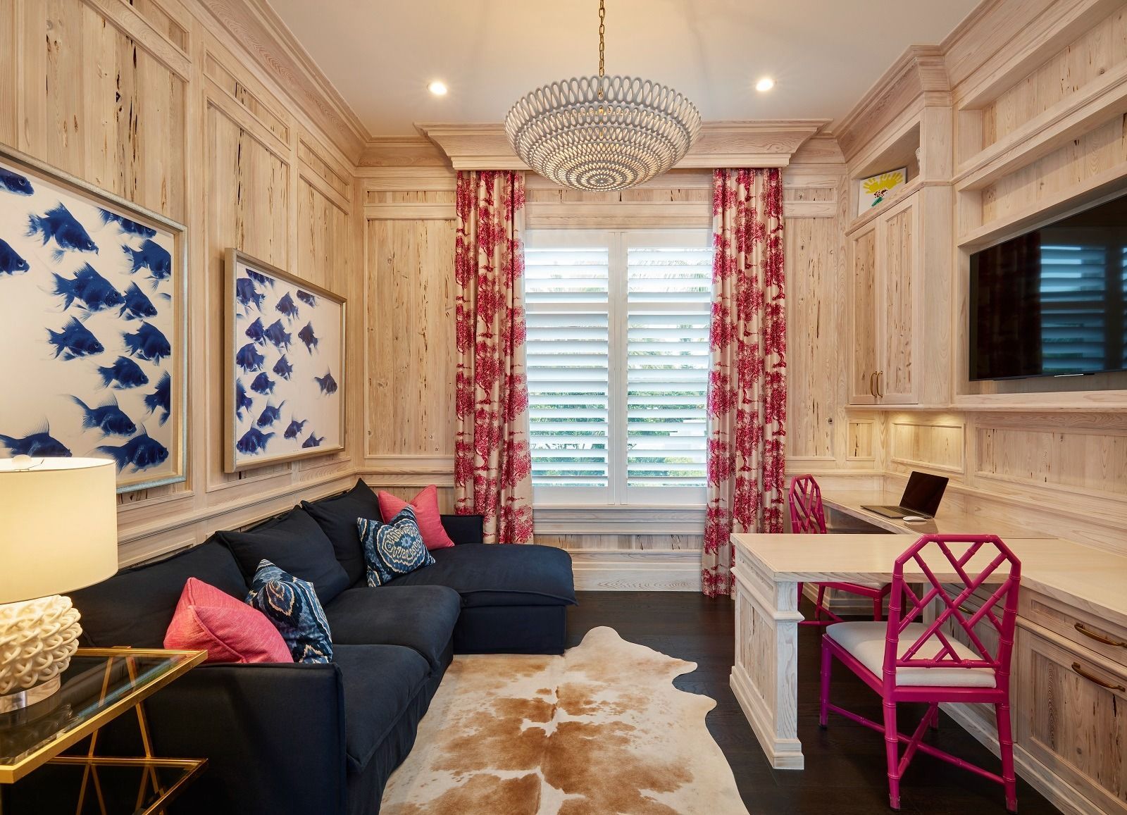 Home office with navy blue sectional, white desk, and pink chair, wood paneling, and patterned curtains.