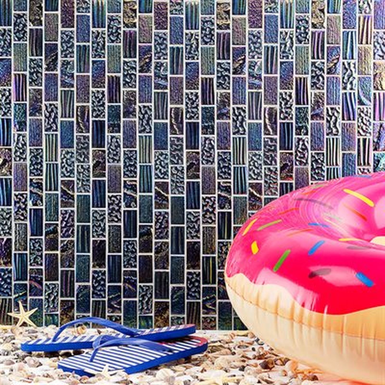 Blue mosaic tile wall with donut float and flip-flops on beach pebbles.