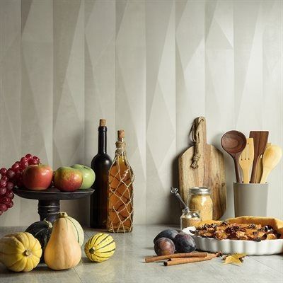 Still life: food and kitchen items on a counter. Pumpkins, apples, pie, bottles, cutting board, and utensils in front of a textured wall.