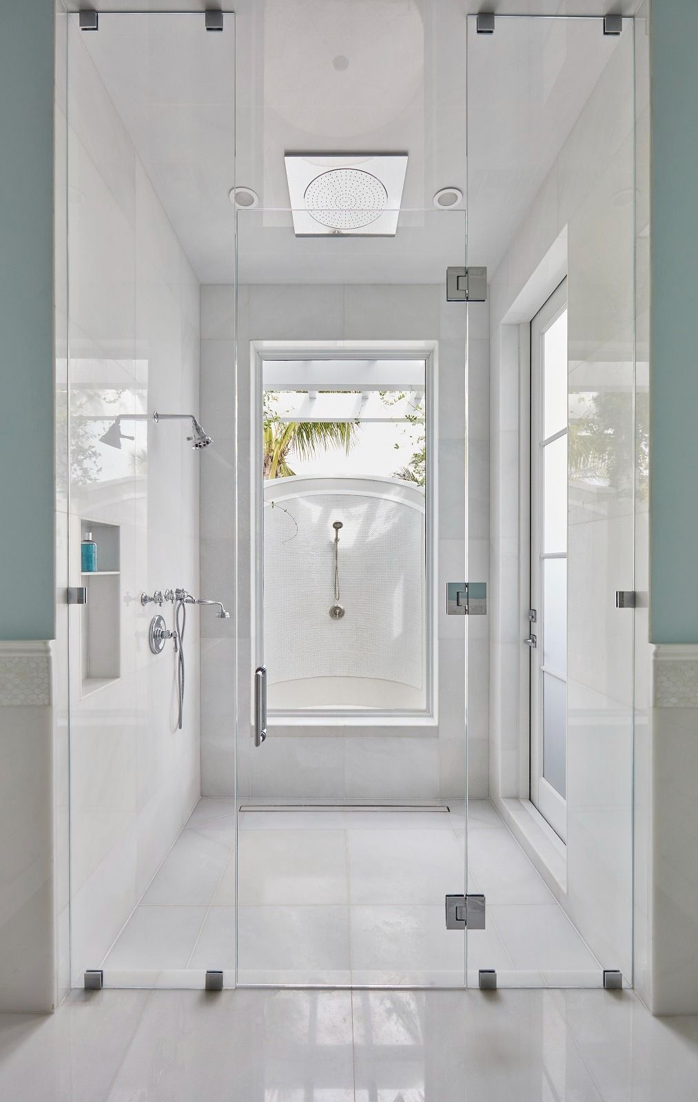 Glass-enclosed shower with white tiled walls and floor, a window at the far end, and a partially open door to the right.