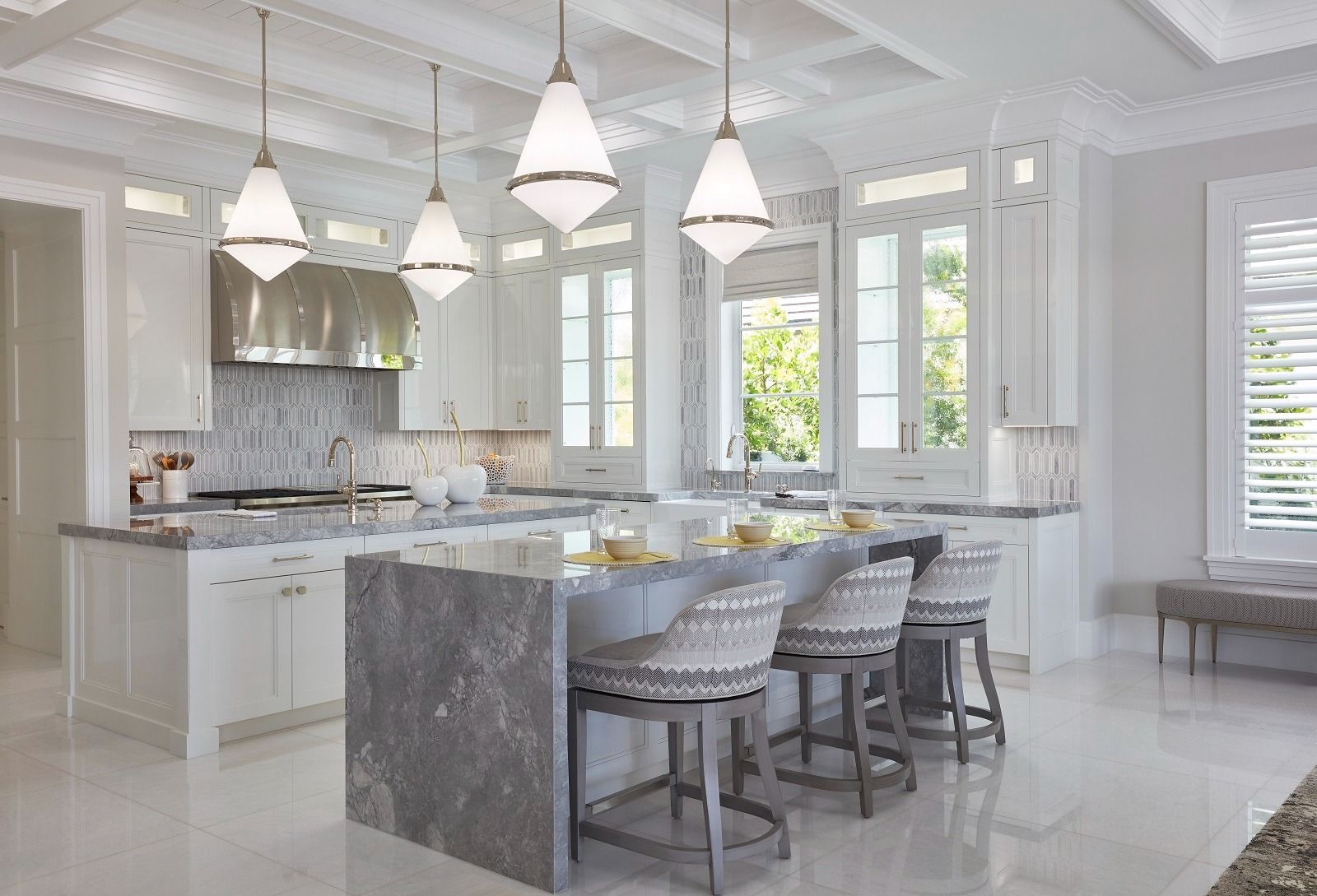 Elegant white kitchen with marble countertops, island with bar stools, pendant lights, and large windows.