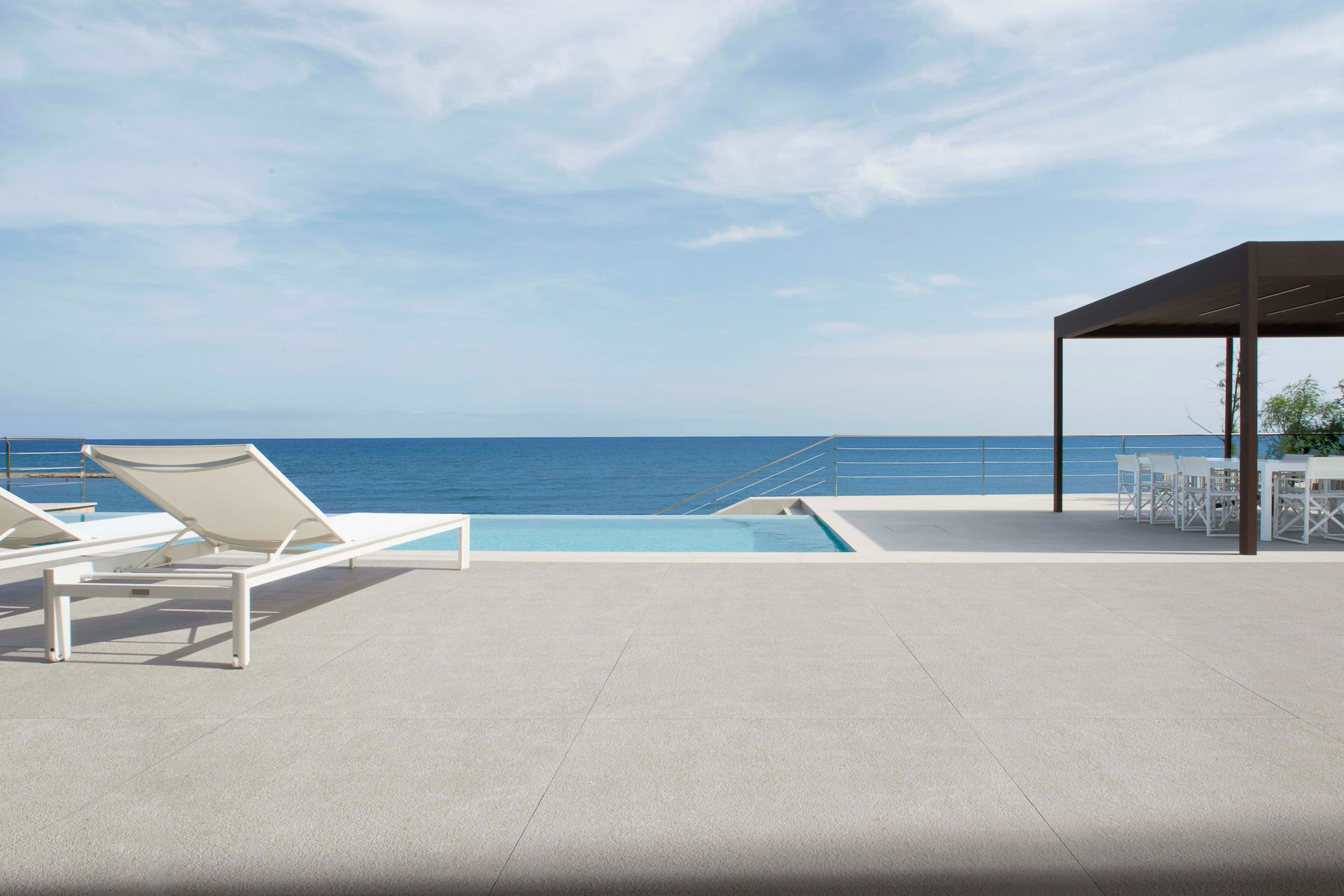 Poolside view of ocean, lounge chairs, pergola with table, blue sky.