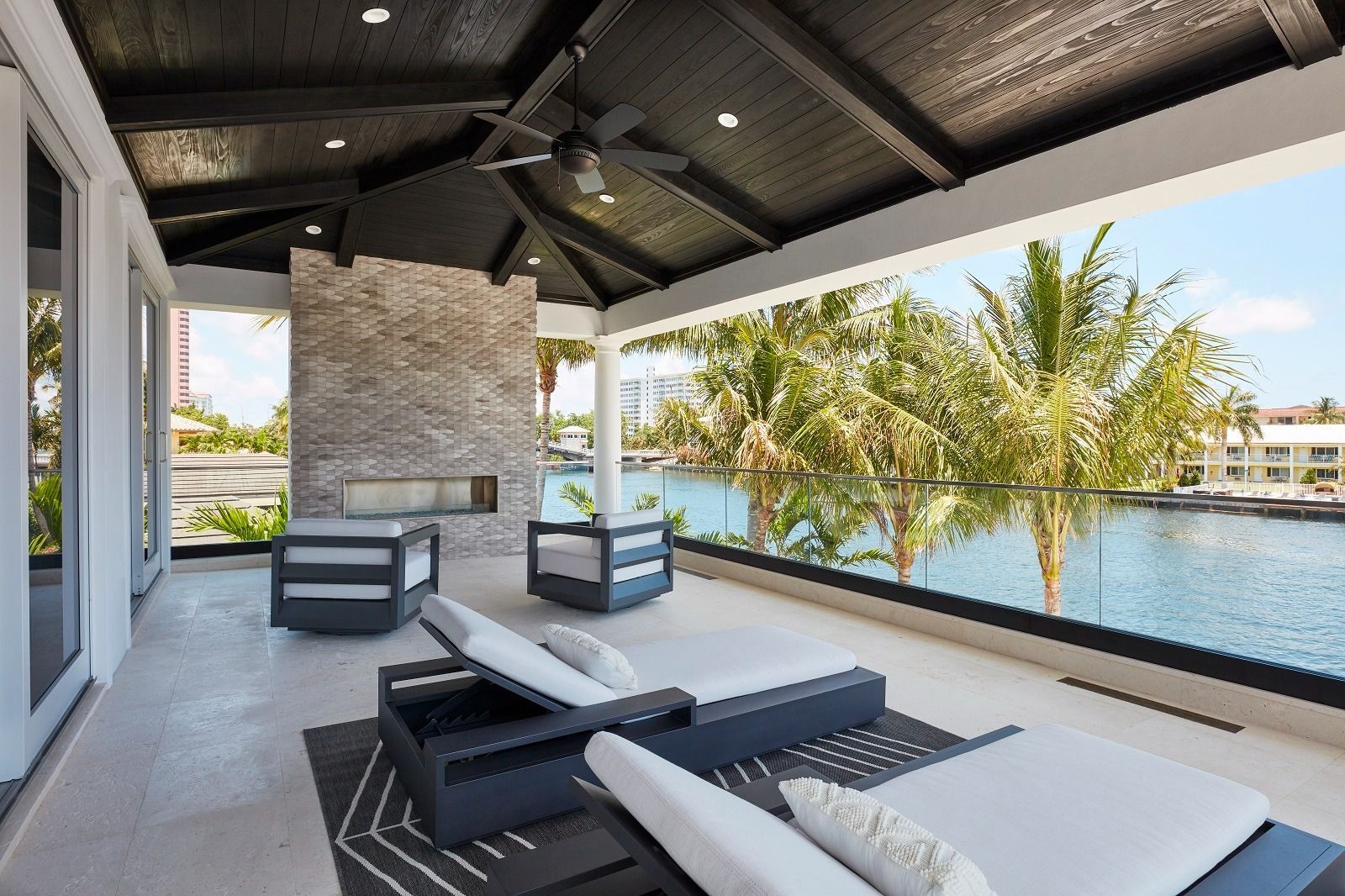 Patio with lounge chairs, fireplace, and view of water and palm trees.
