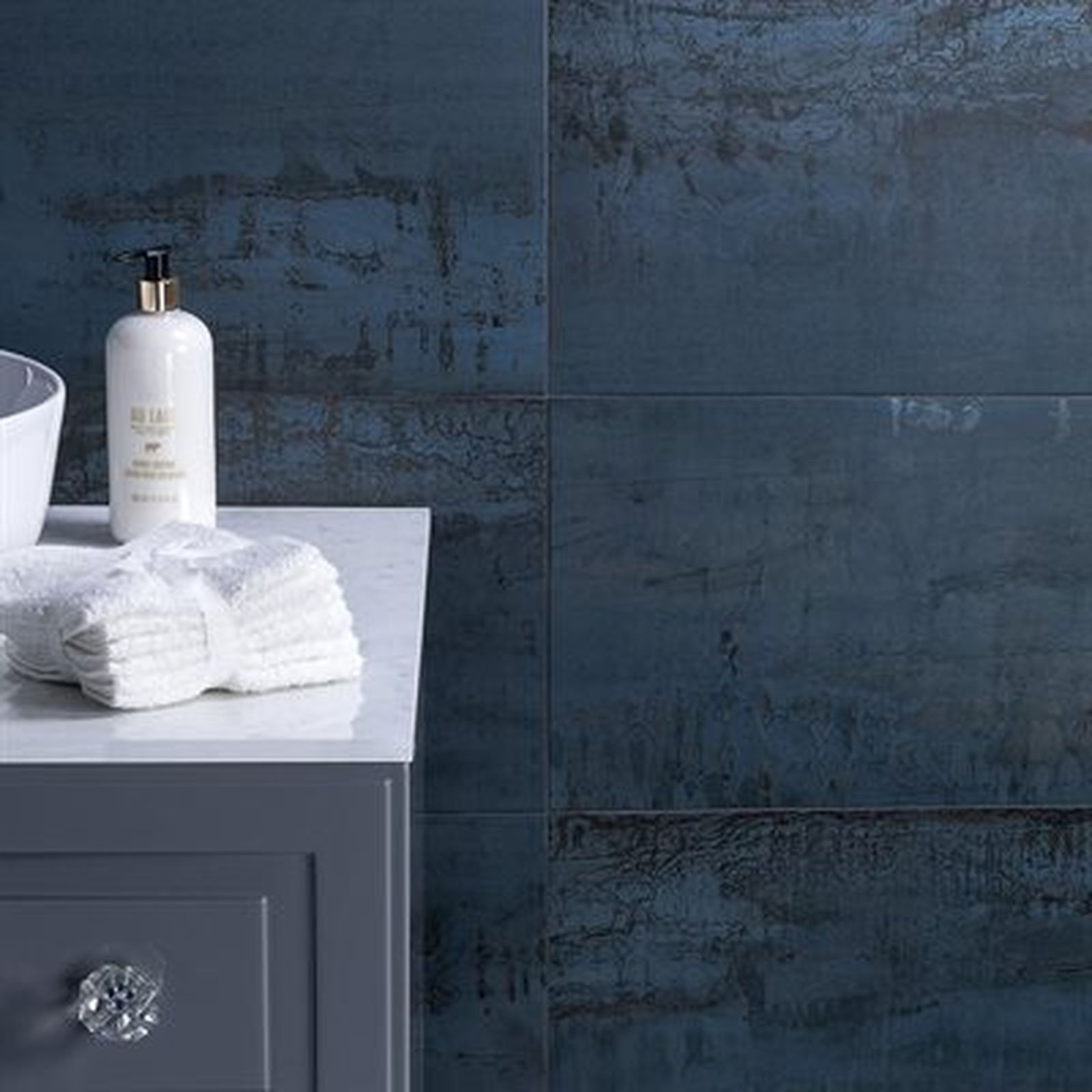 Bathroom with blue textured tiles, marble countertop, towels, soap dispenser, and gray cabinet.