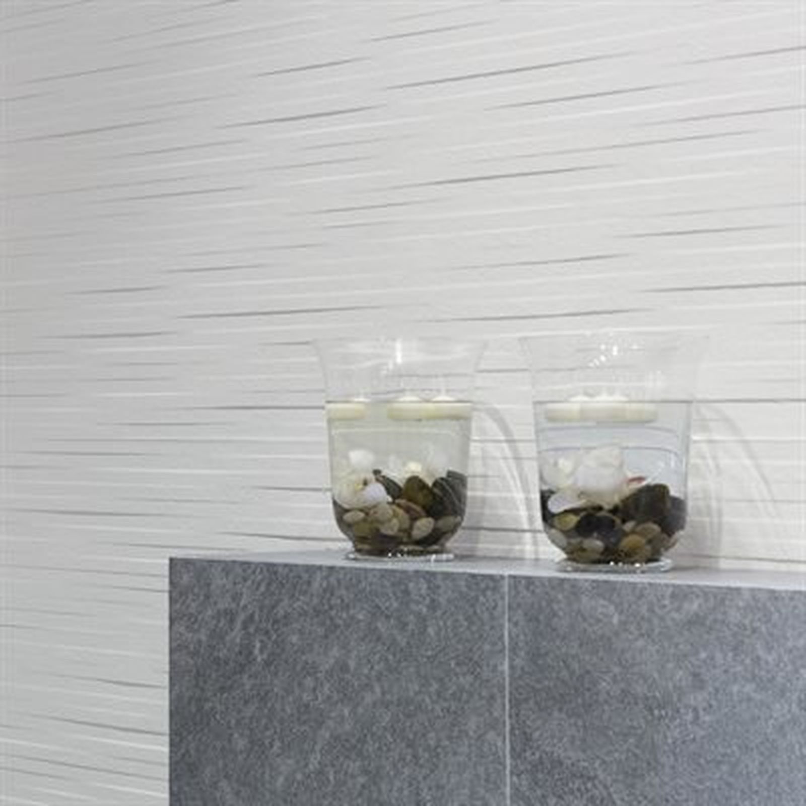 Two clear glasses with pebbles and floating objects on a gray stone shelf, white textured wall backdrop.