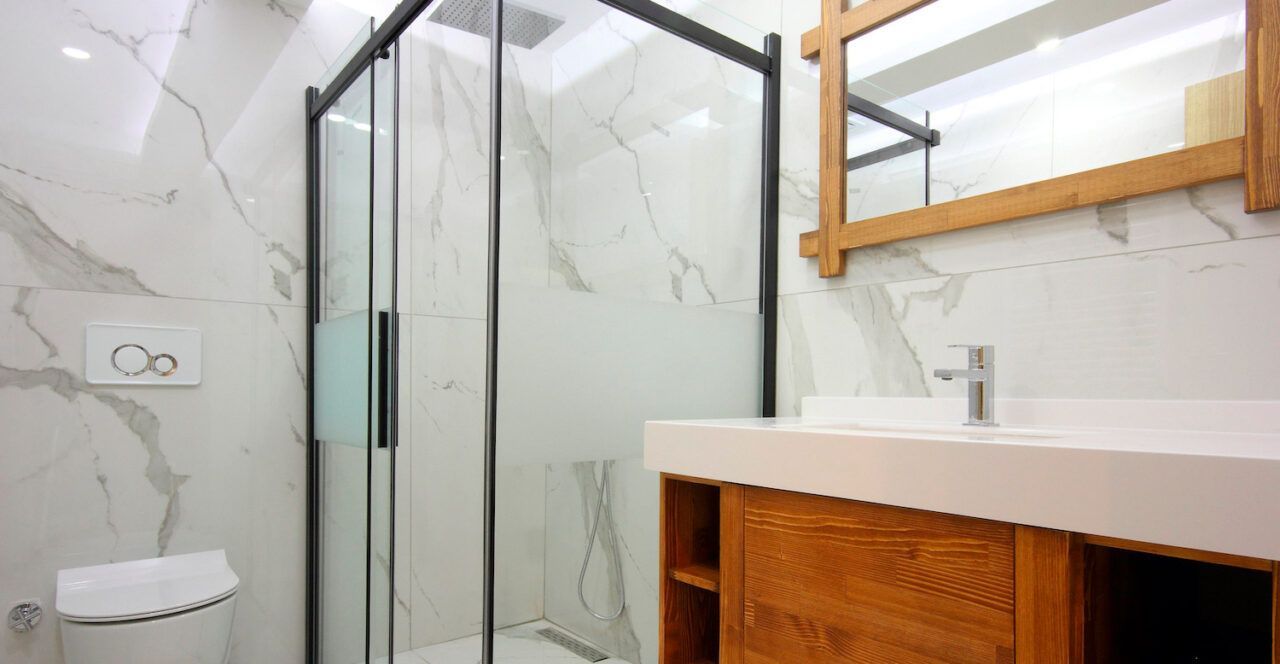 Bathroom with marble walls, glass shower, wooden vanity, and square mirror.
