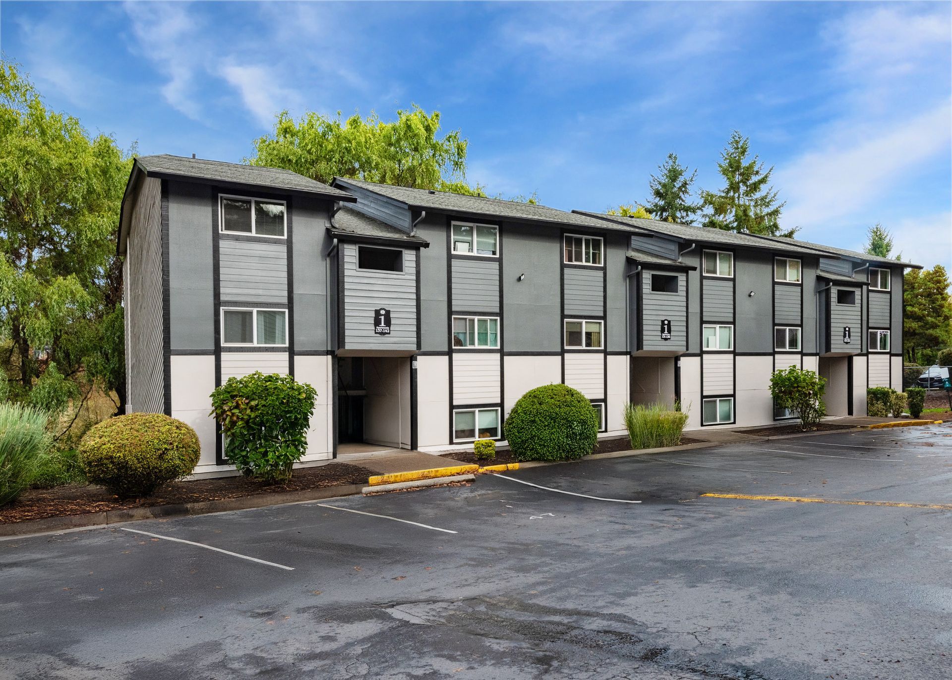 Exterior view of a gray and white apartment building with landscaping and a parking lot.