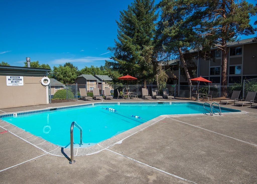 Outdoor community pool with lounge chairs and red umbrellas beside apartment buildings.