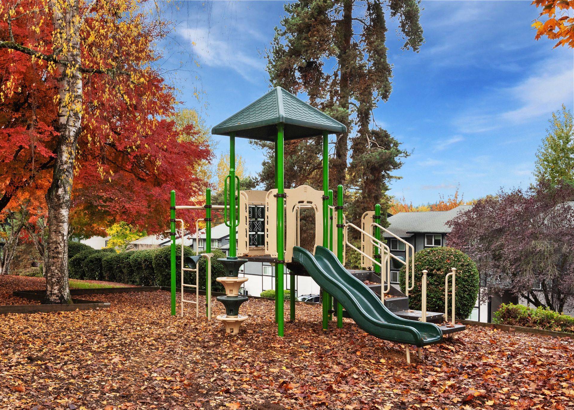 Playground with a green play structure, slide, and autumn leaves in a residential community.