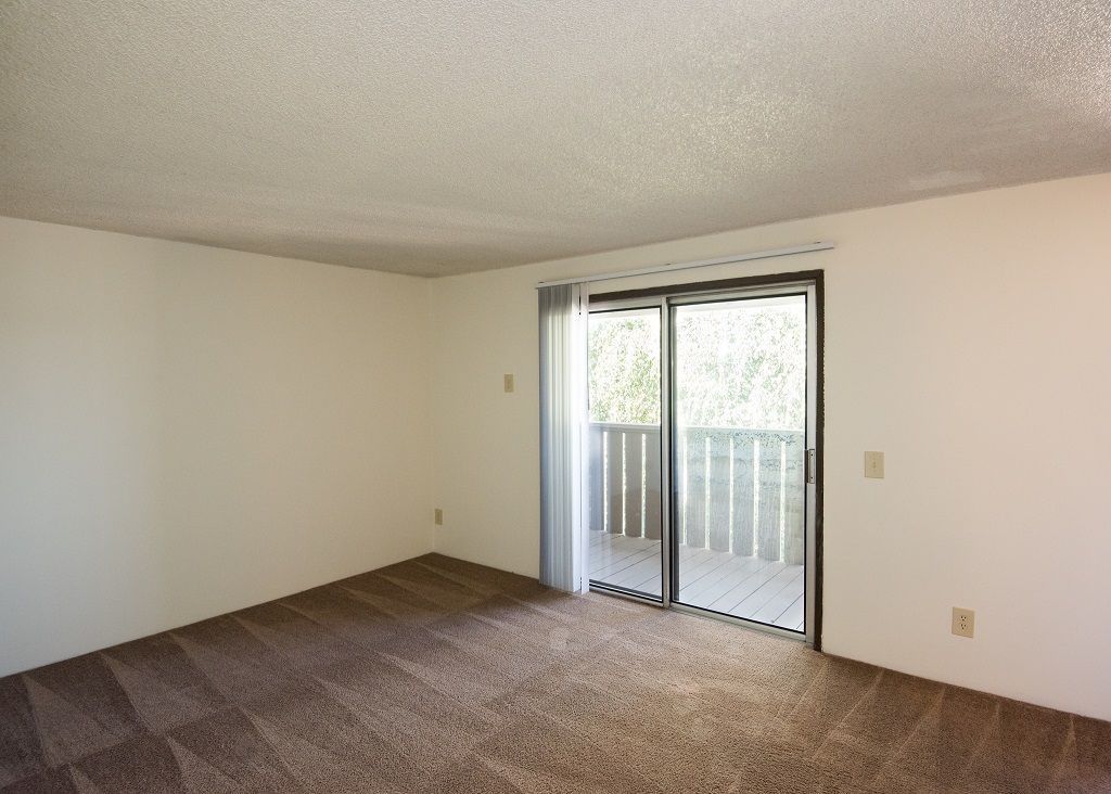 Empty living room with sliding glass door to balcony and beige carpet.