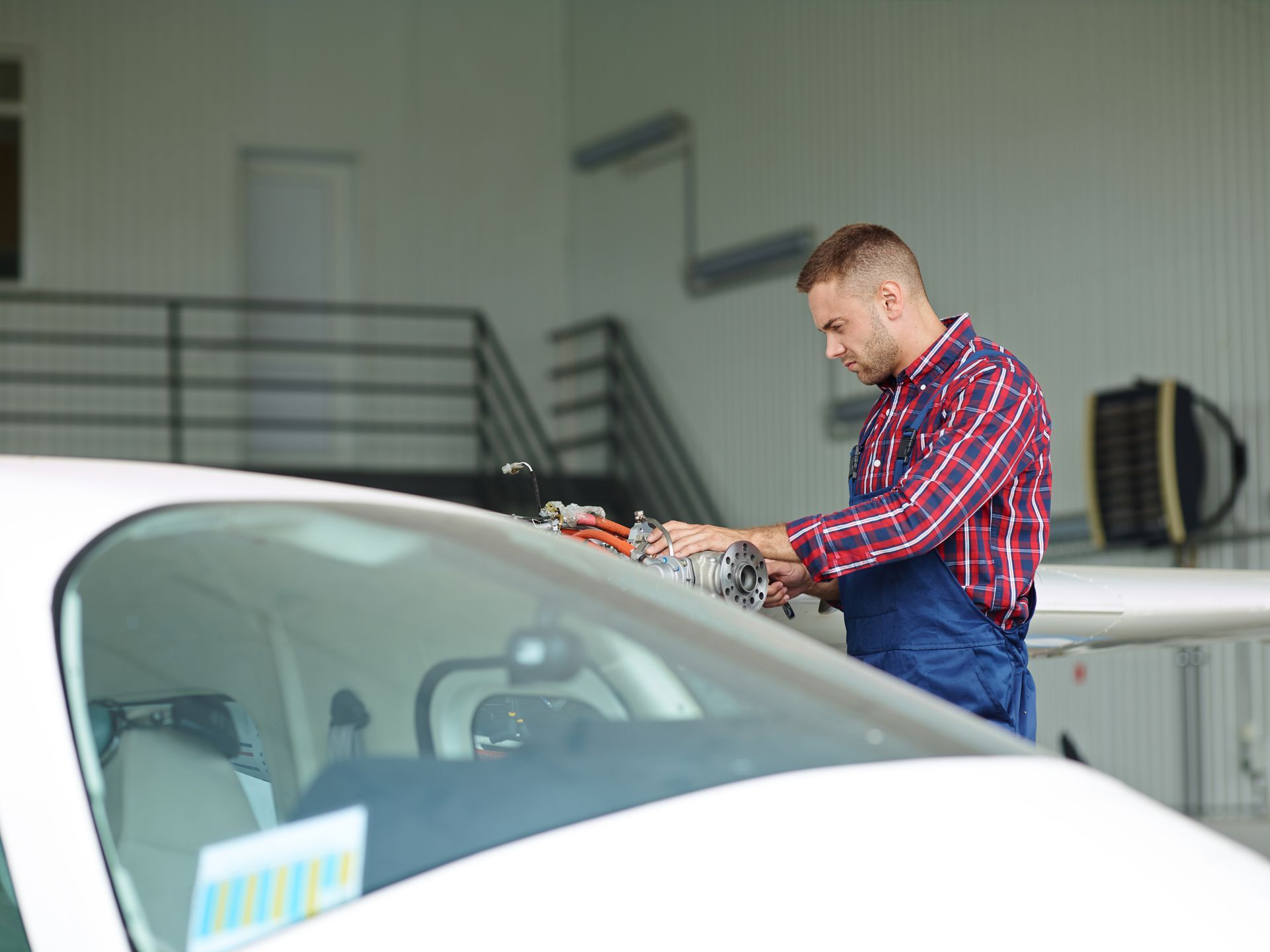 A technician in a red plaid shirt and blue overalls inspects the engine of a small aircraft inside a hangar.