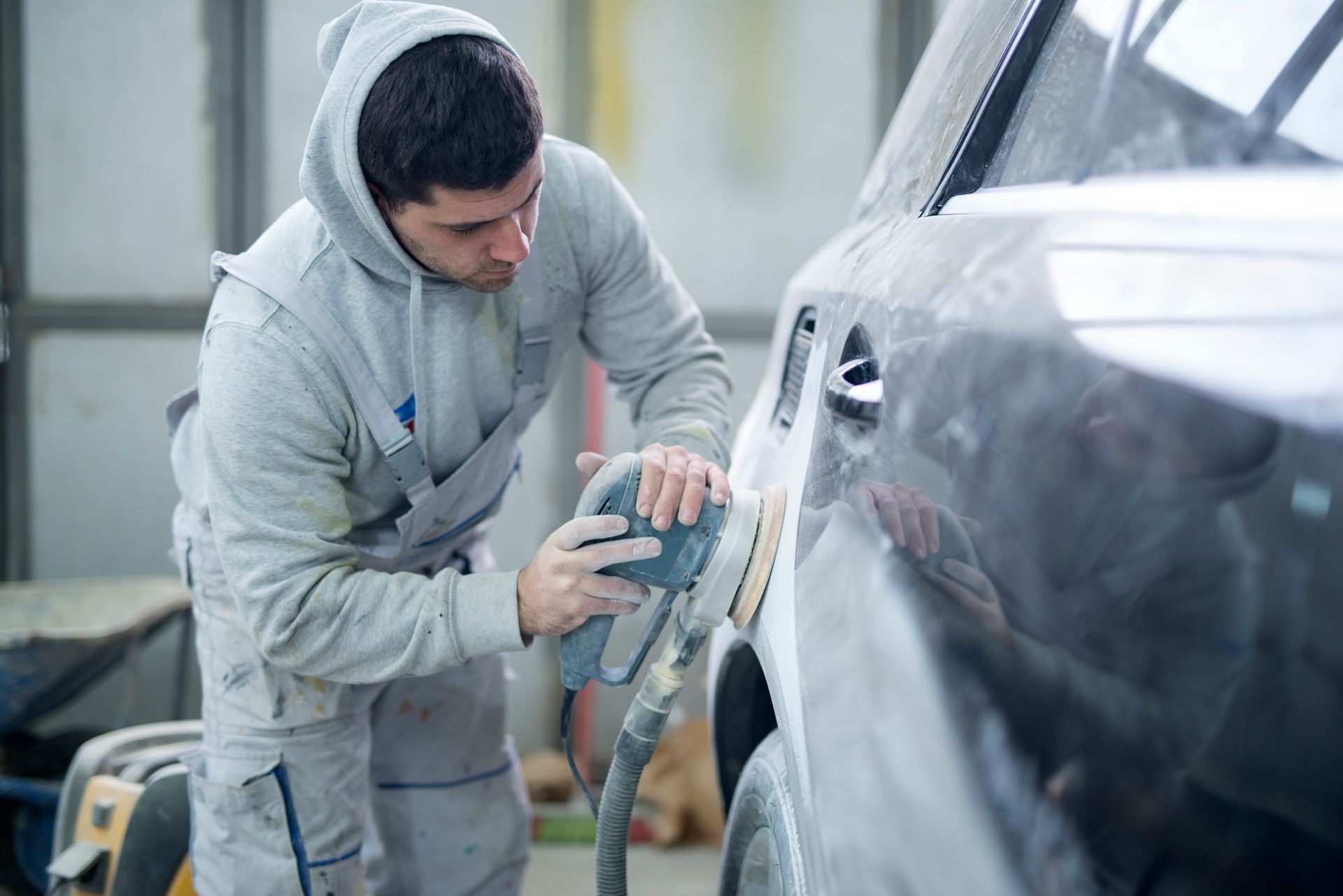 A person in a gray hooded coverall uses a power sander on the side of a car in a workshop.