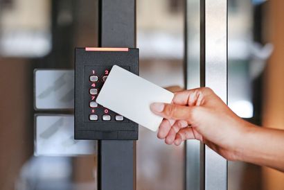 A person uses a smart card to scan into an electronic wall-mounted reader against a gray concrete background.