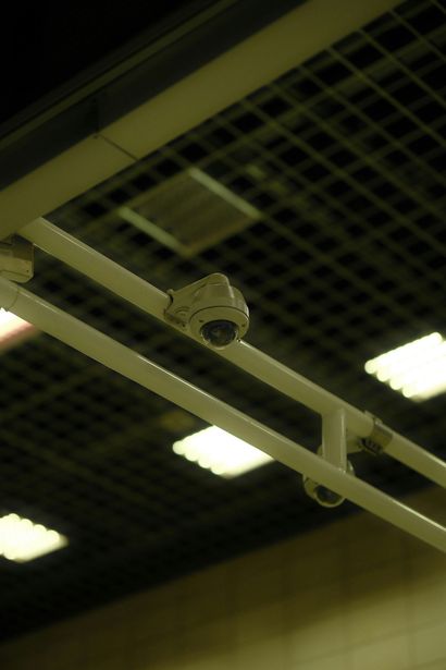 A security camera mounted on a white metal frame structure against a dark ceiling with light panels.