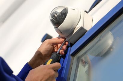 A person in a blue uniform uses a screwdriver to install or repair a dome-shaped security camera on a wall.