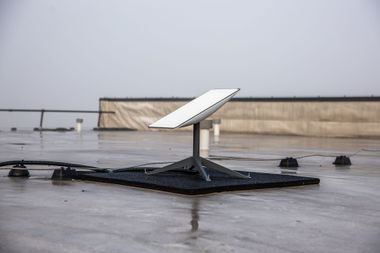 A Starlink satellite dish mounted on a black, weighted base sits on a wet, flat rooftop under a gray, overcast sky.