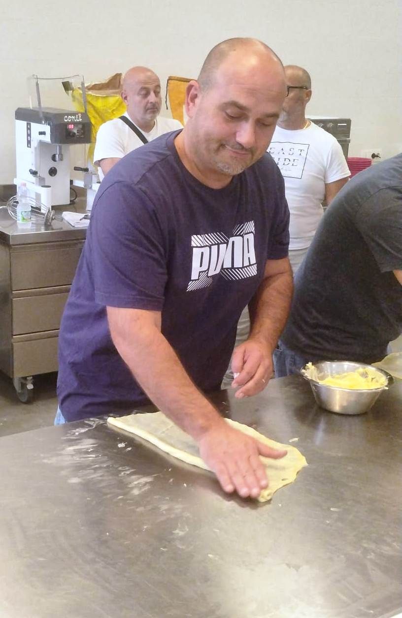 Un uomo con una maglietta blu della Puma stende la pasta su un bancone in acciaio inossidabile; altri lo guardano in cucina.
