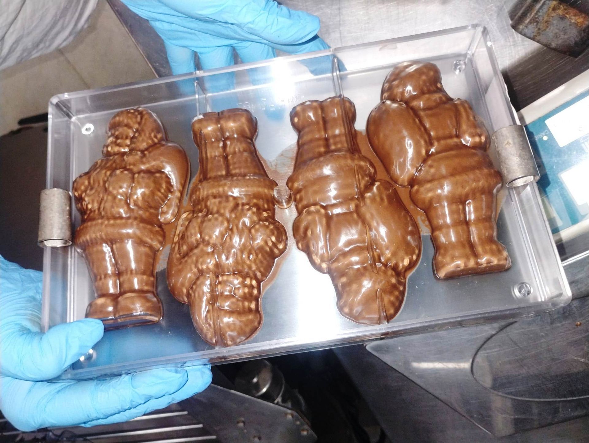 Quattro figure di Babbo Natale di cioccolato in un vassoio di plastica trasparente, tenute da mani guantate.