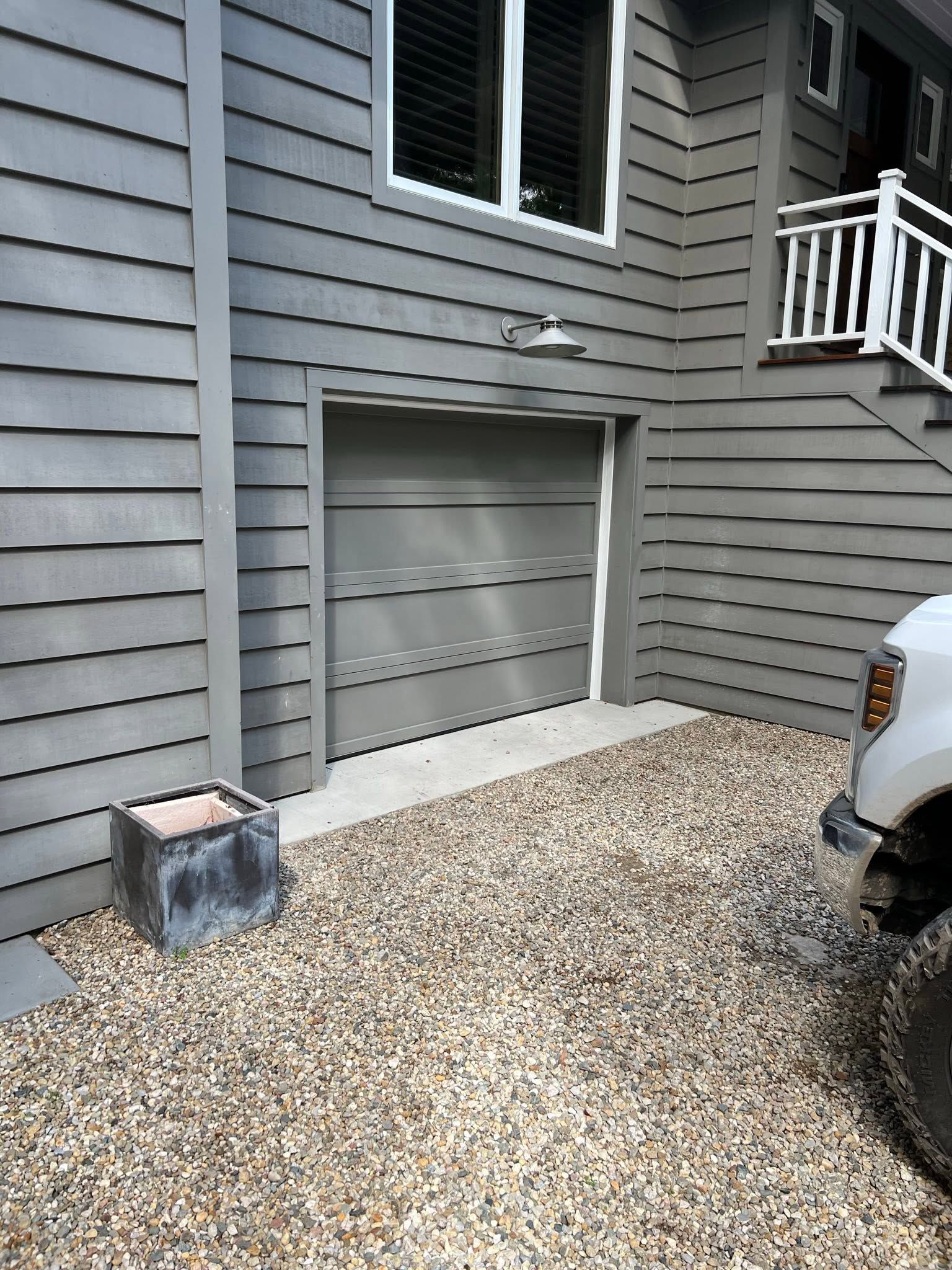 Gray house siding with a small basement door, gravel driveway, and part of a parked car on the right.