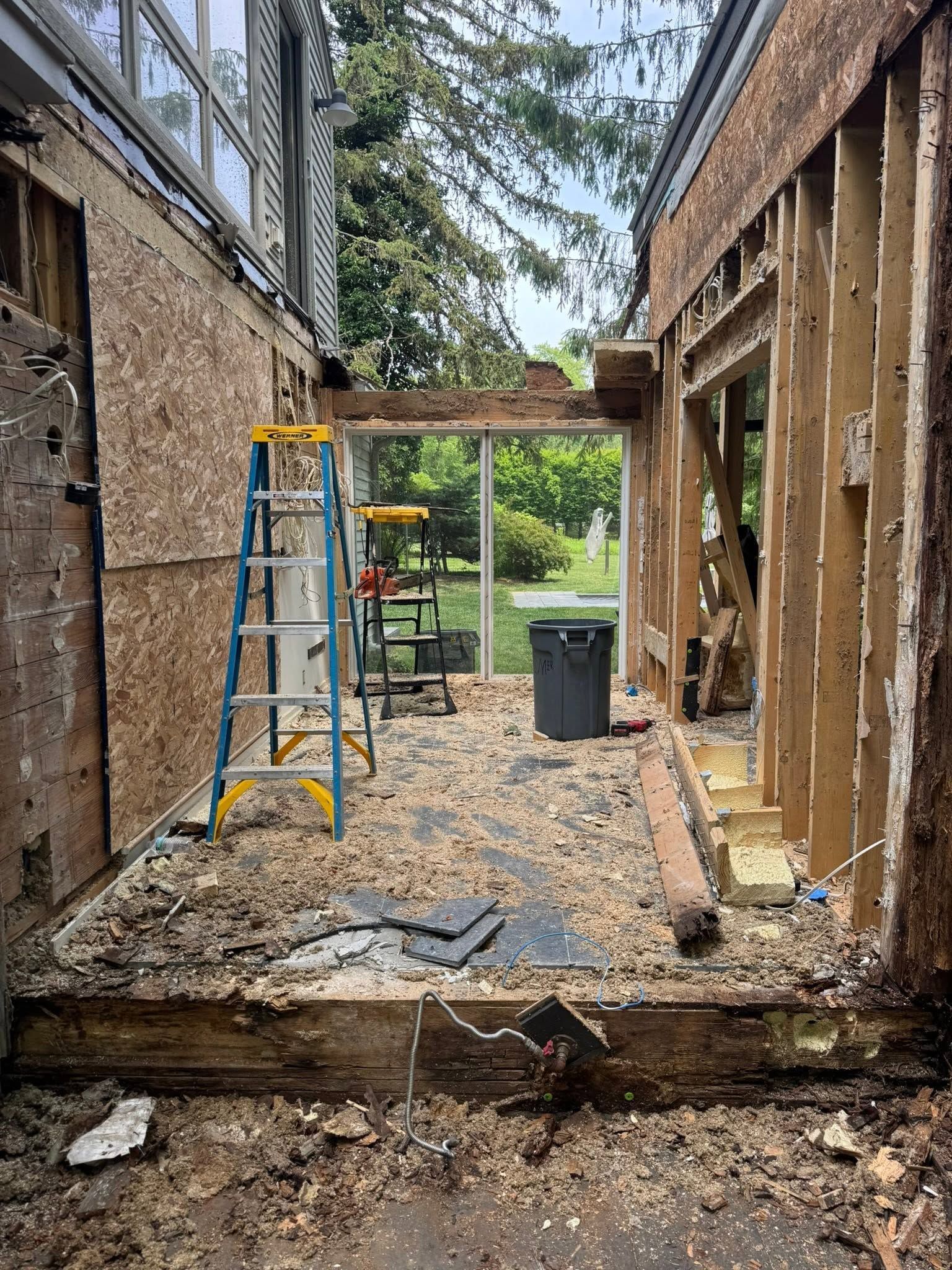 Partially demolished room with exposed studs, ladder, and debris opening to a backyard