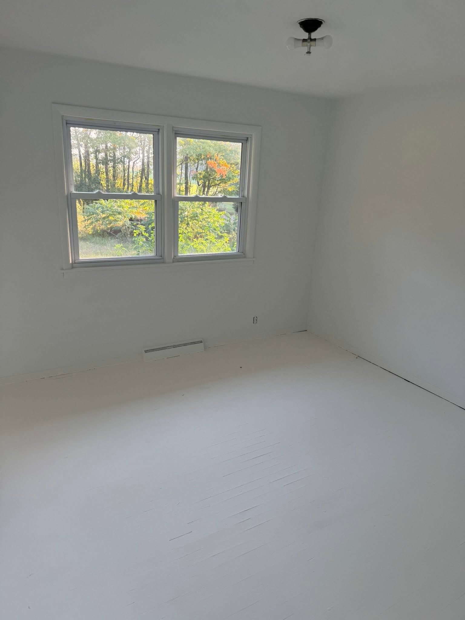 Empty white room with a window and bare light fixture, lit by daylight.