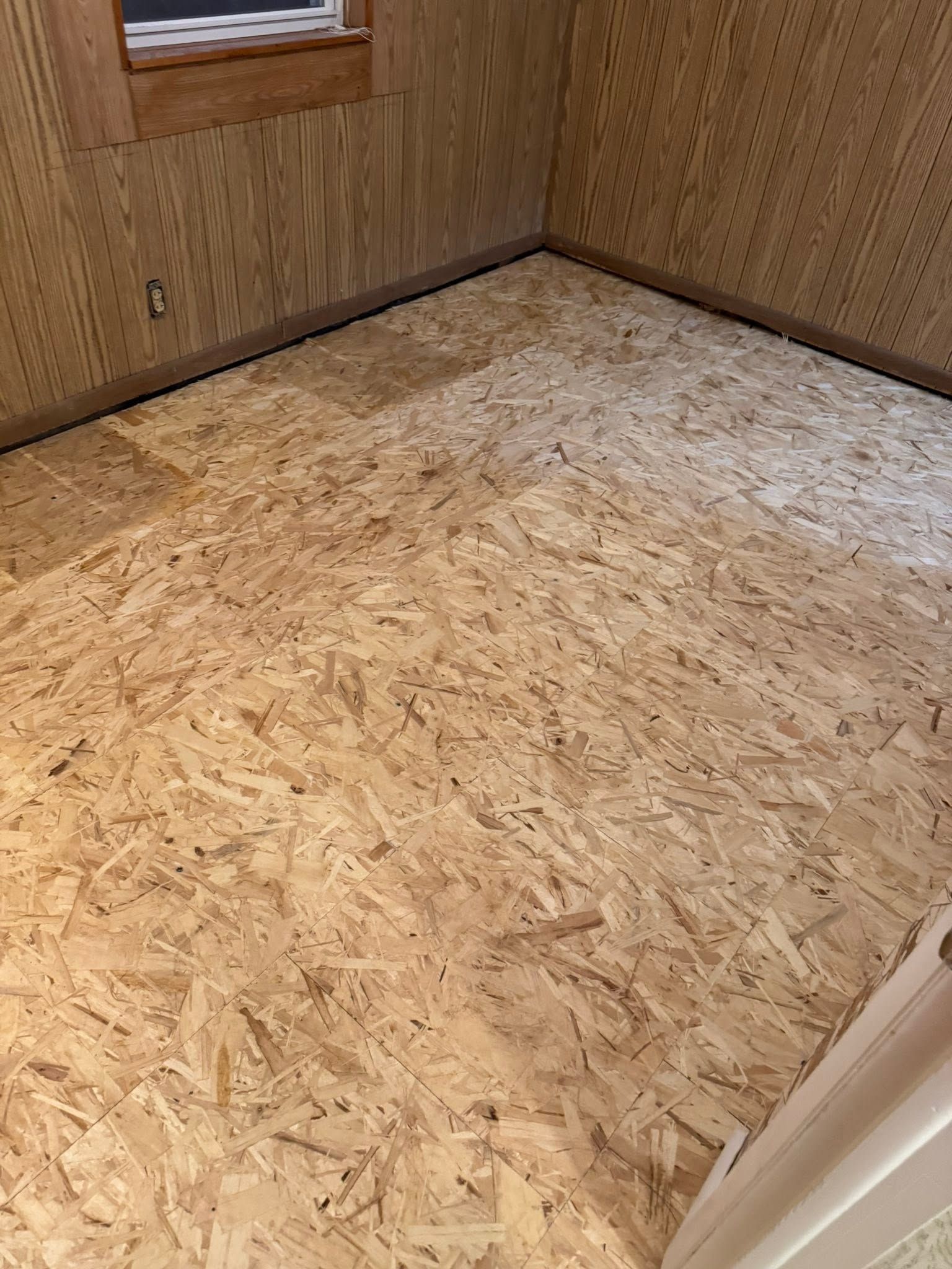 Empty room with plywood subfloor, wood-paneled walls, and a small window.