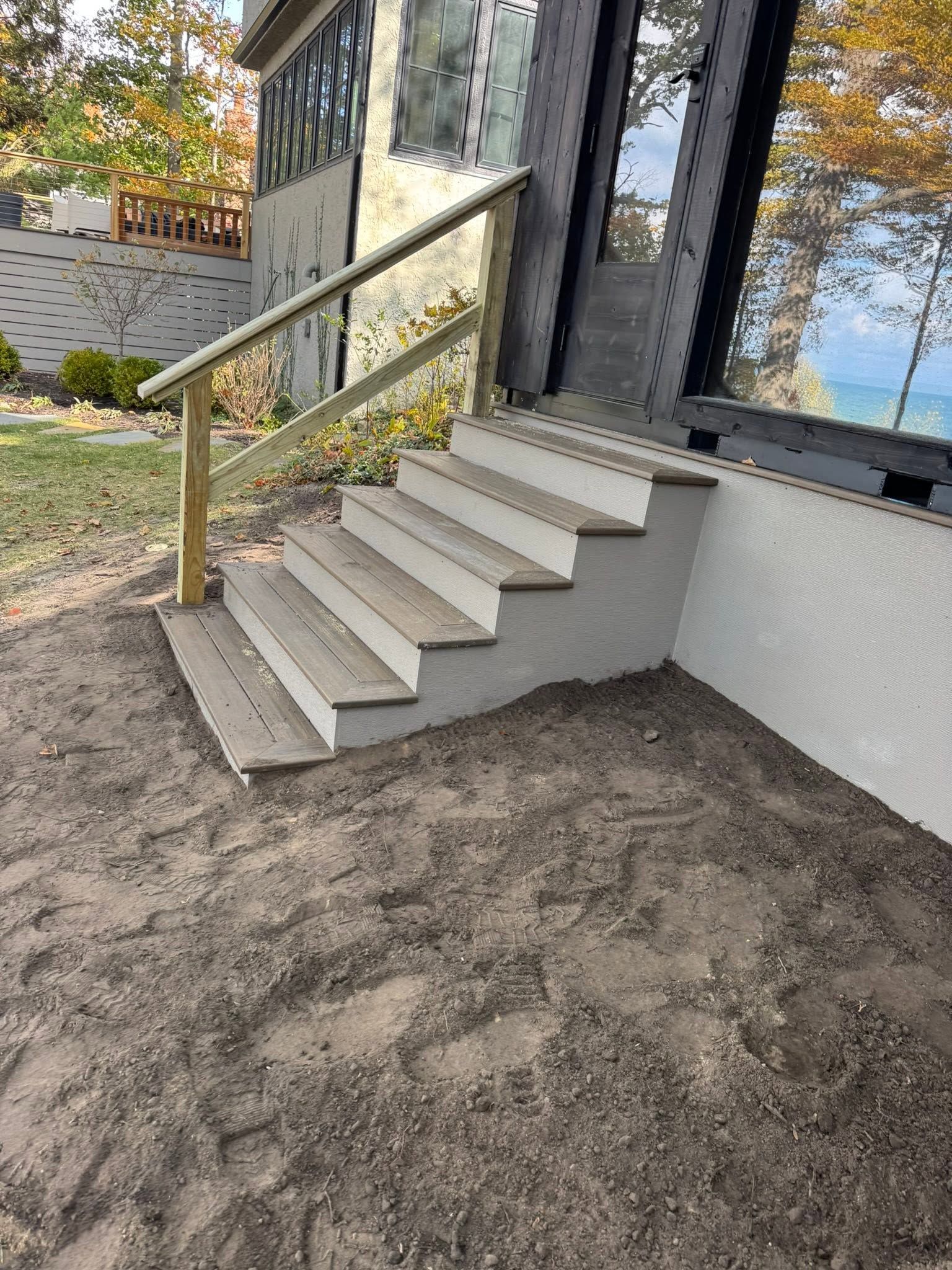 Exterior stairs with beige handrail beside a dark house wall and gravel ground on a sunny day