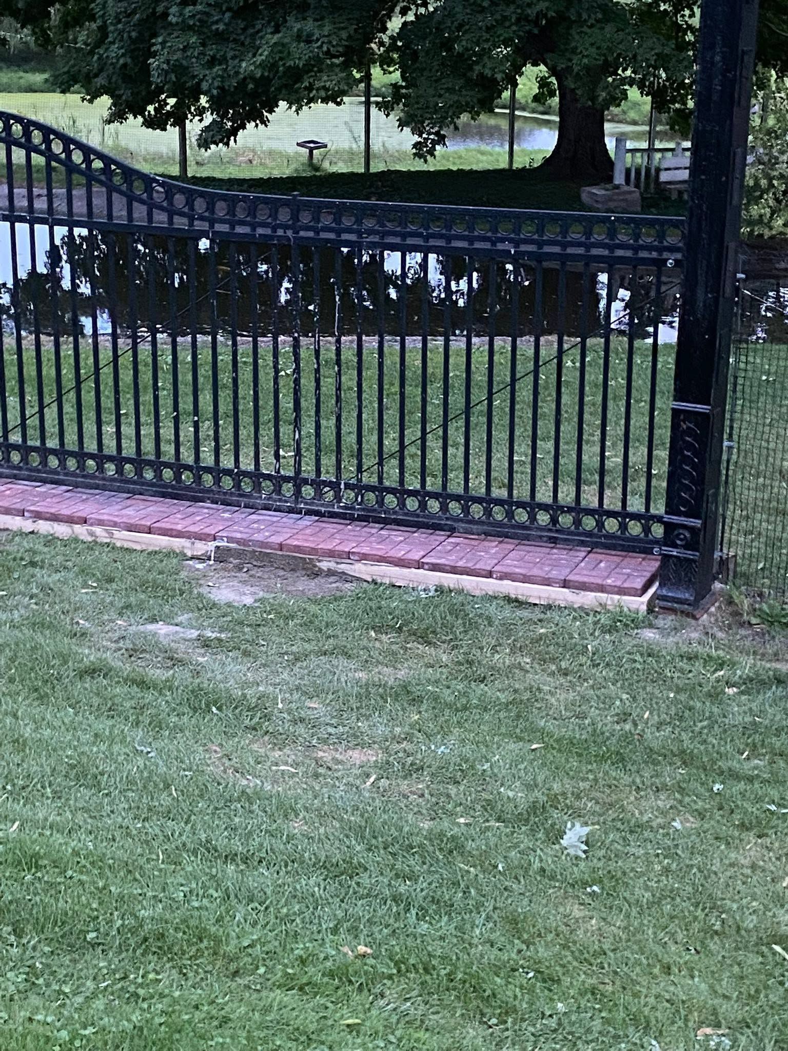 Black iron fence beside a grassy lawn and small pond with trees in the background.