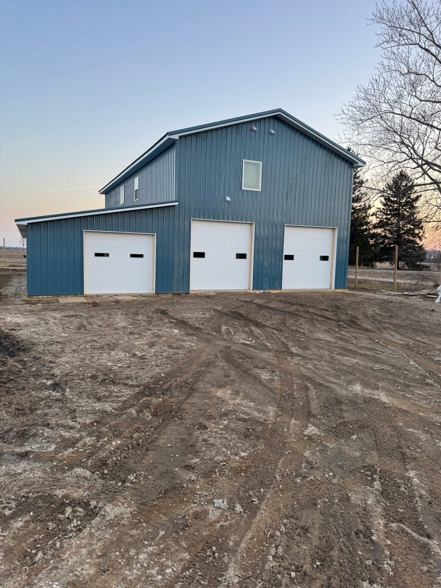 Blue metal barn with three white garage doors on a dirt lot at dusk