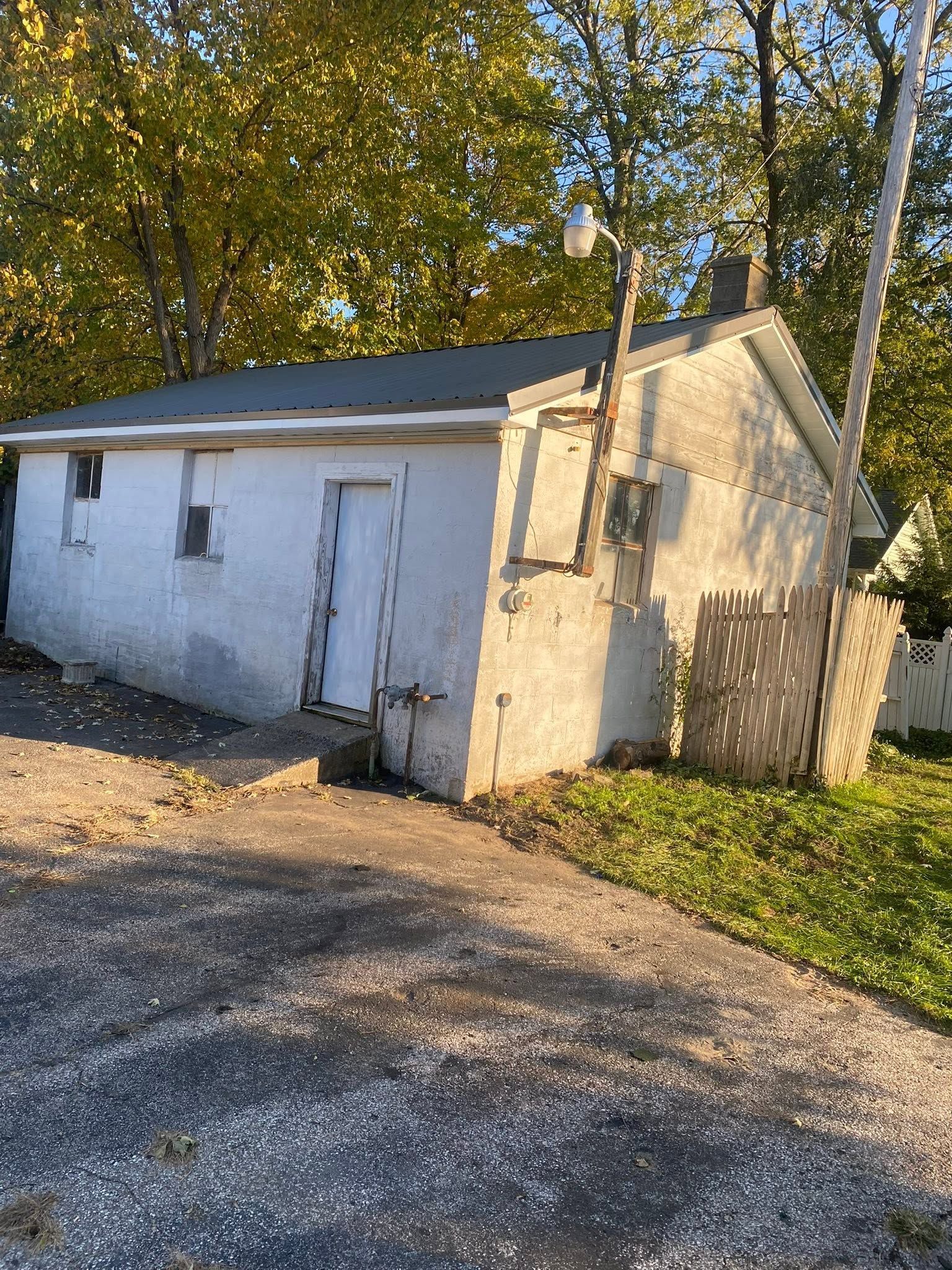 Small white outbuilding with a sloped roof beside a gravel drive and trees in warm sunlight
