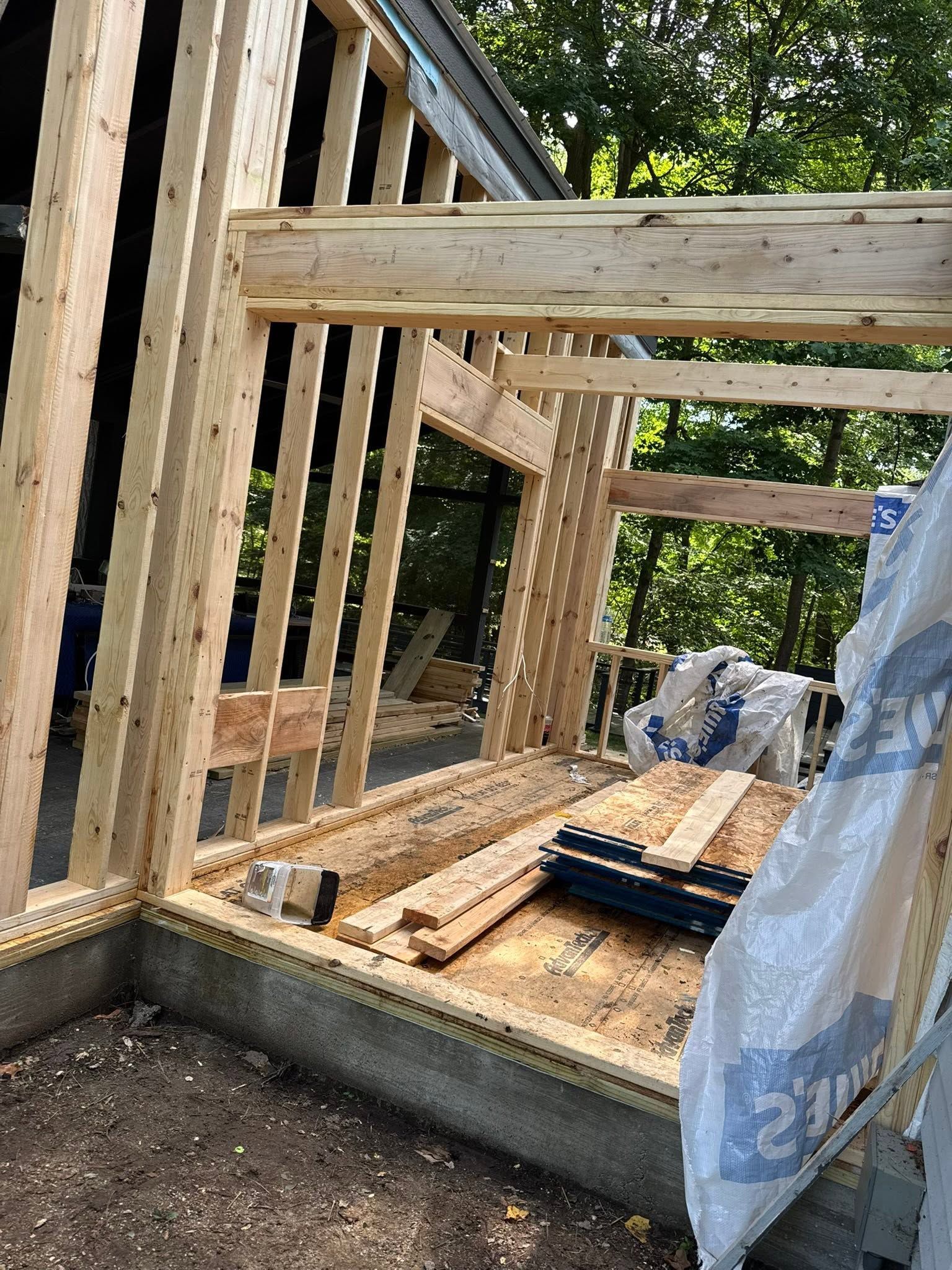 Wood-framed deck under construction beside a concrete foundation, with lumber stacked on the floor.