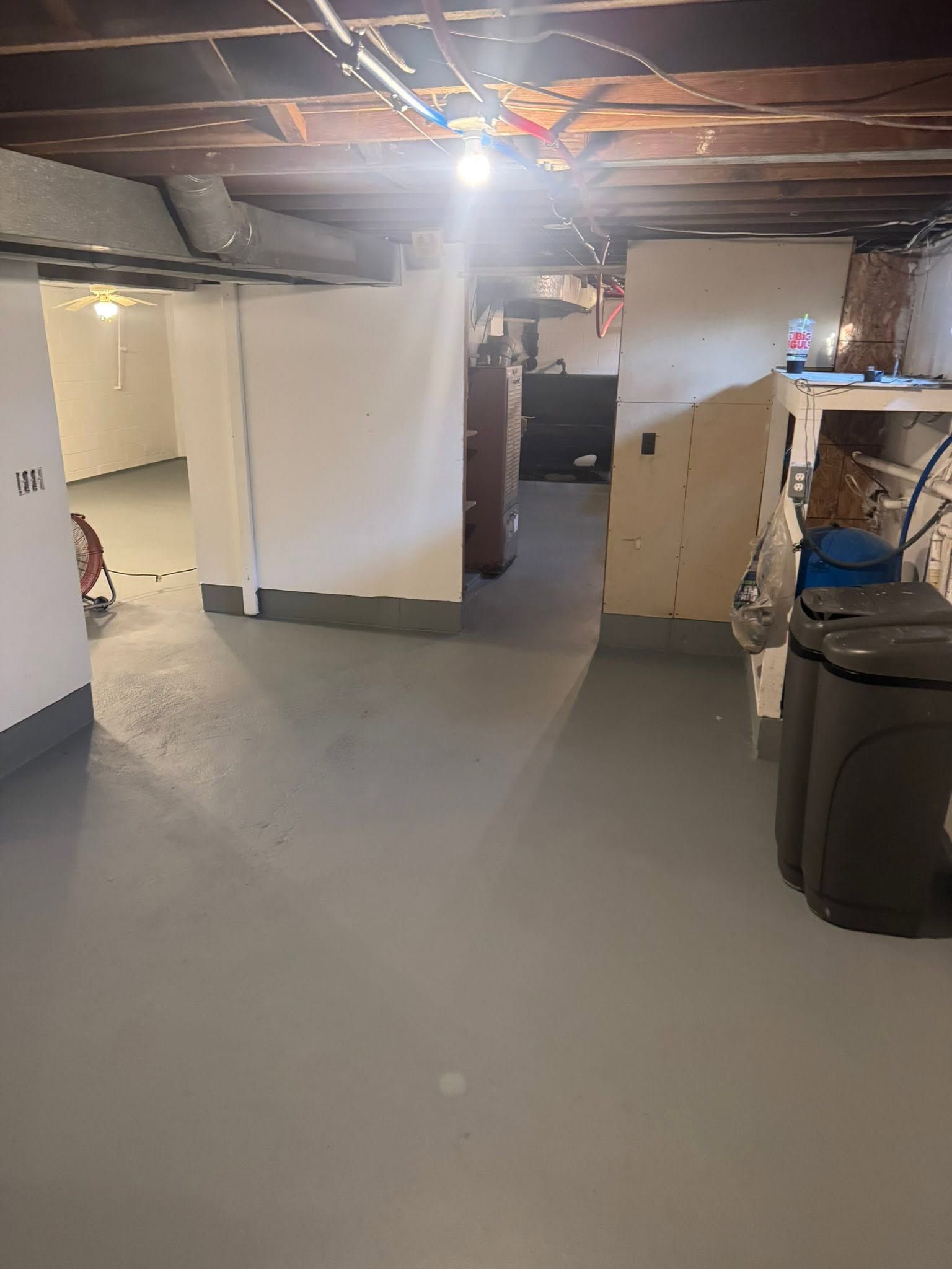 Empty basement with concrete floor, exposed ceiling beams, doorway, storage cabinet, and a trash bin.