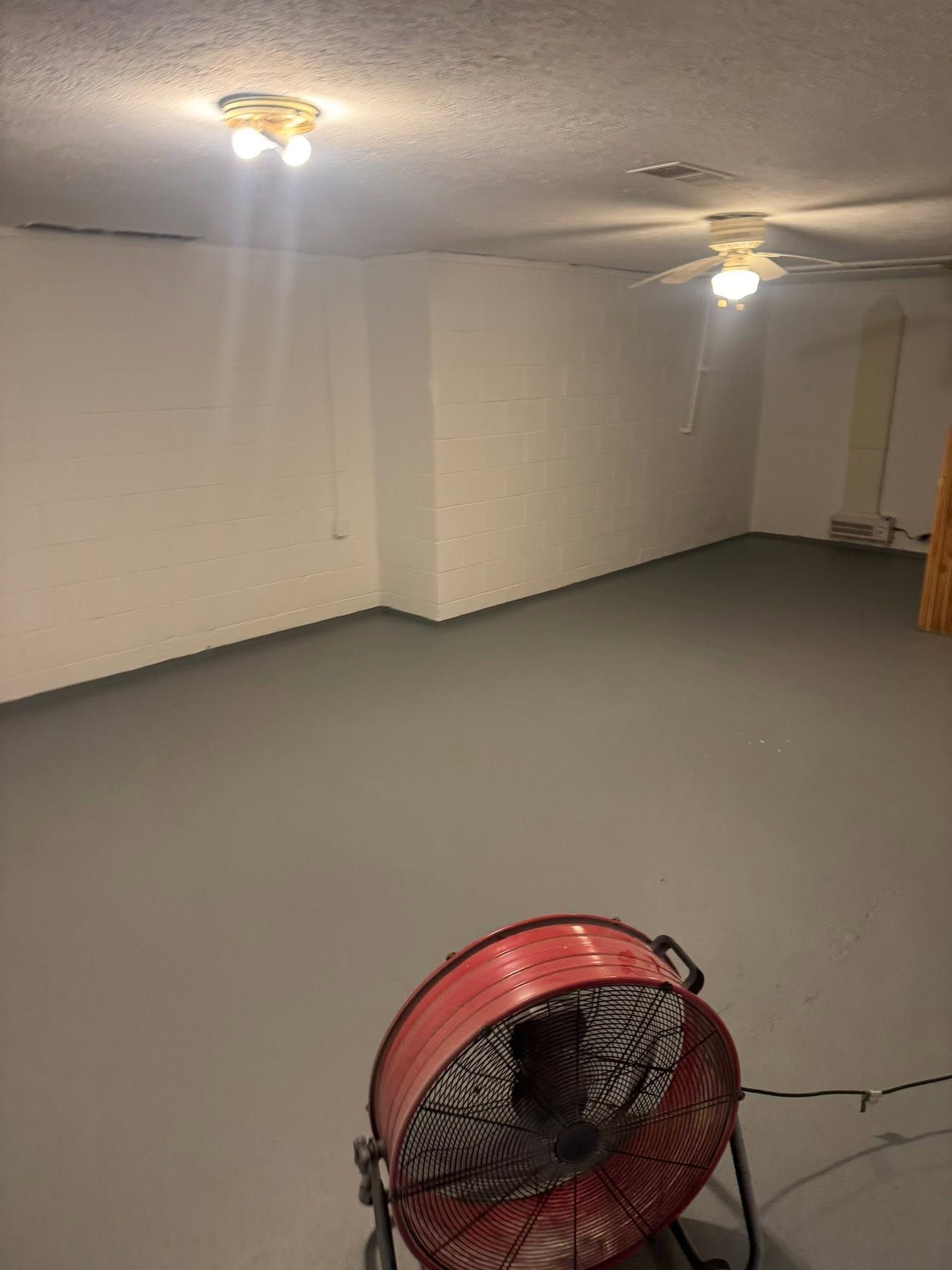 Empty basement with gray floor, white walls, ceiling lights, and a red floor fan in the foreground
