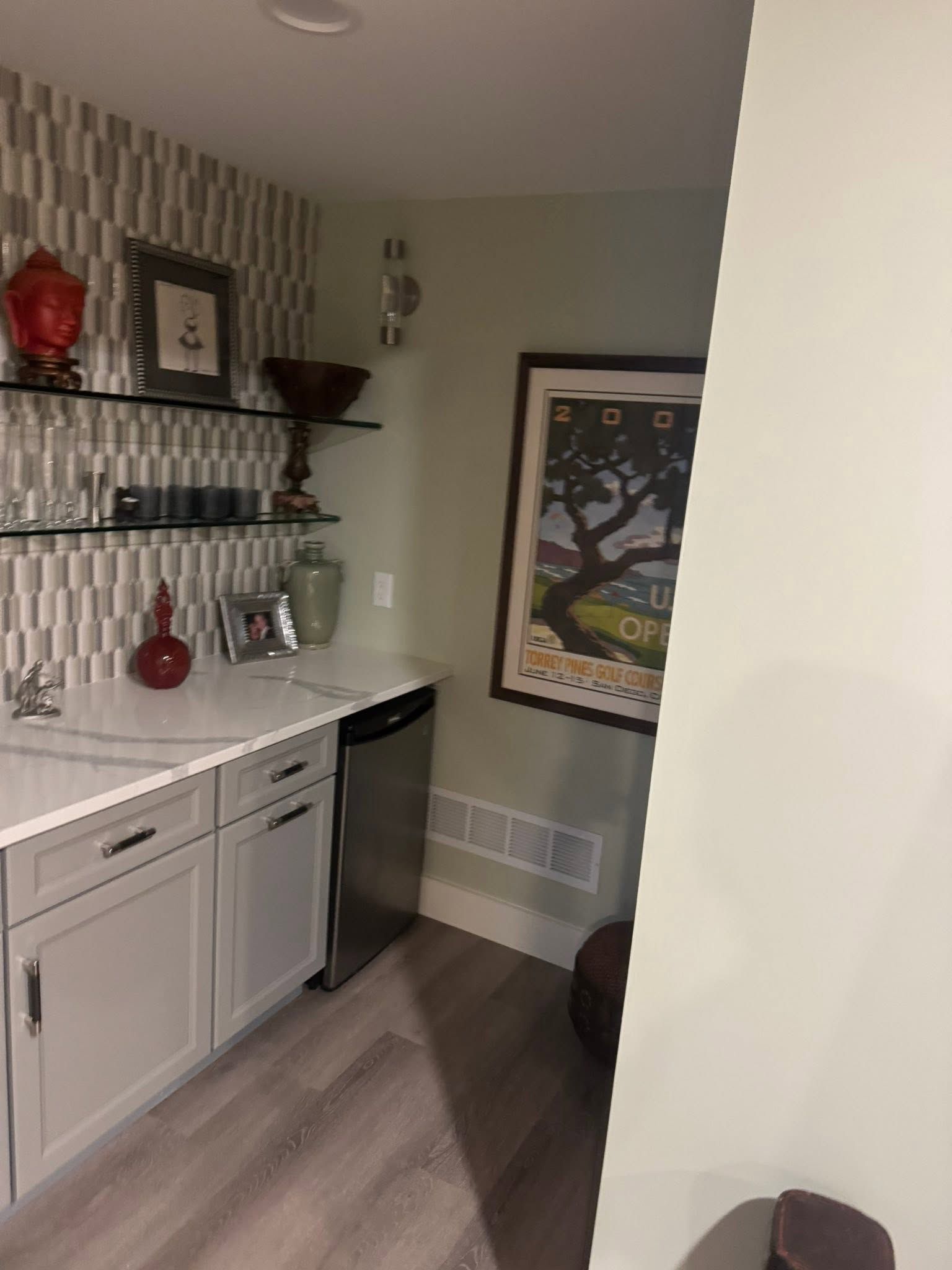 Small kitchen nook with white cabinets, shelves, framed art, and a narrow fridge against a striped wall.
